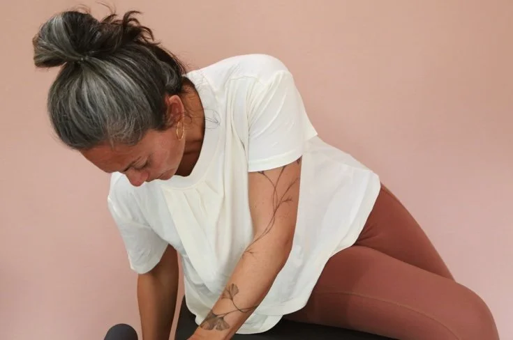 A woman with gray hair in a bun, wearing a white shirt and brown pants, leaning forward with her head down, against a light pink wall.