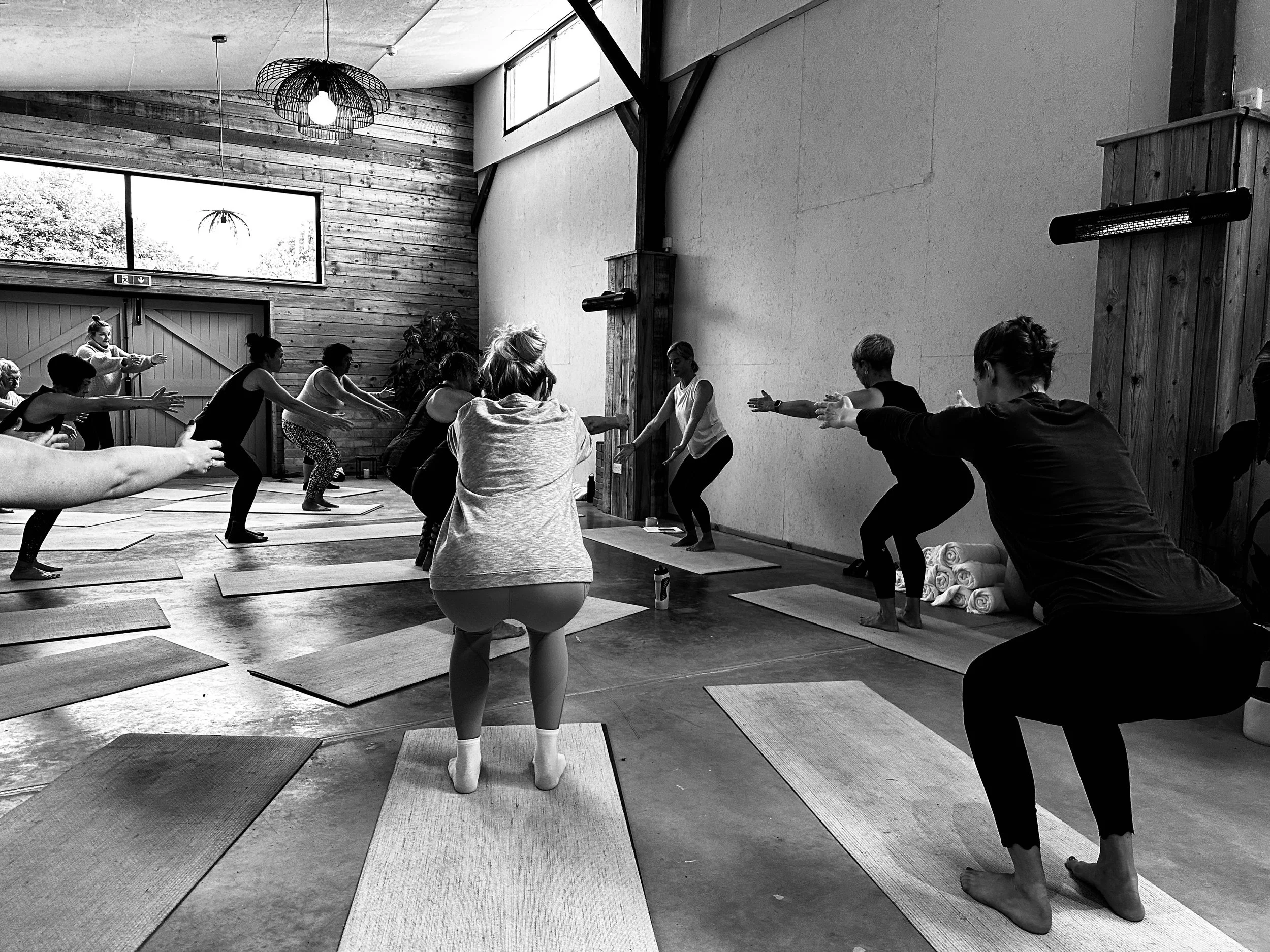 People participating in a yoga class in a studio, some are stretching or in squatting poses, with a instructor leading the class at the front.