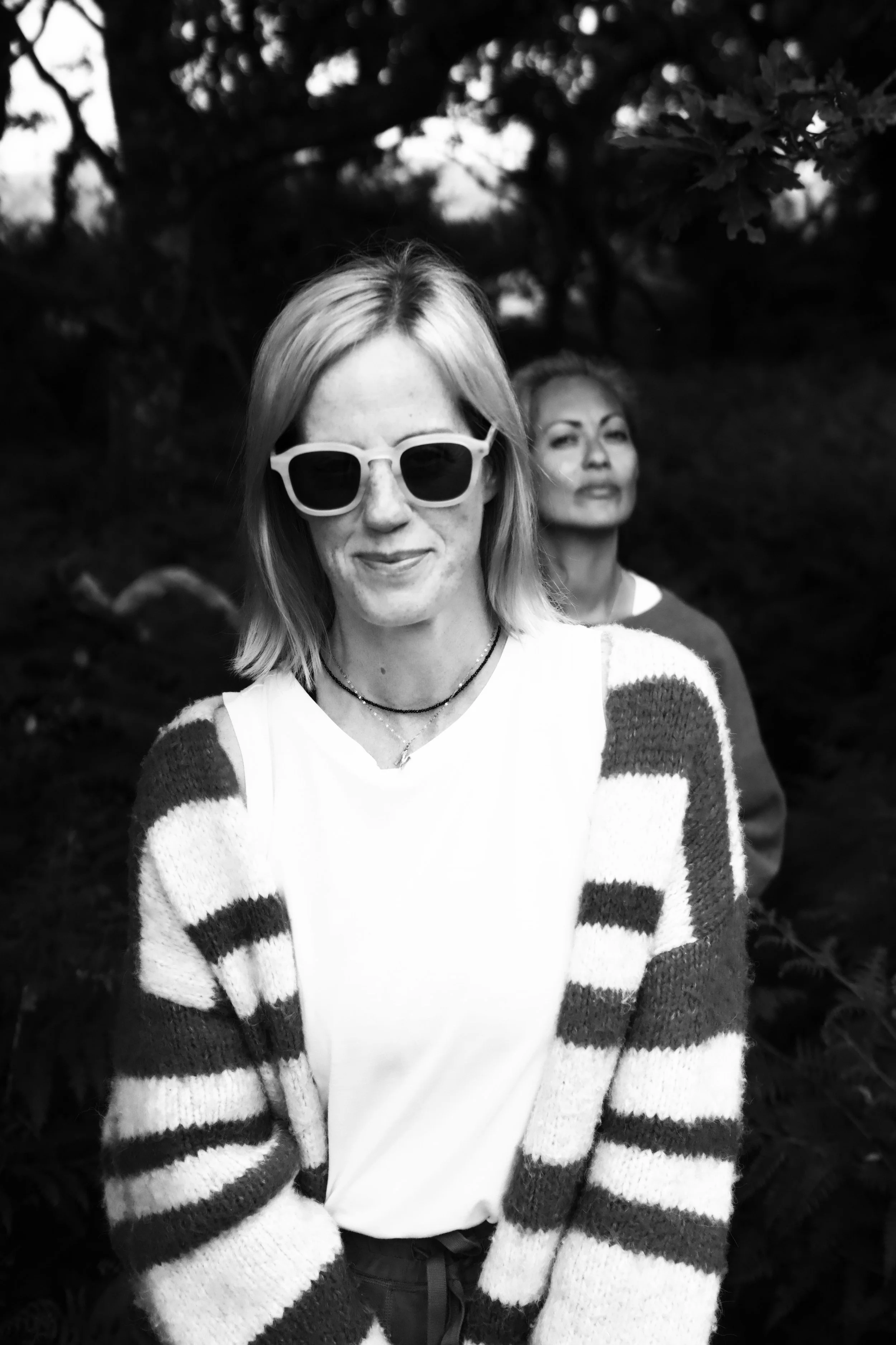 Two women outdoors, one in the foreground wearing glasses and a striped sweater, and another in the background.