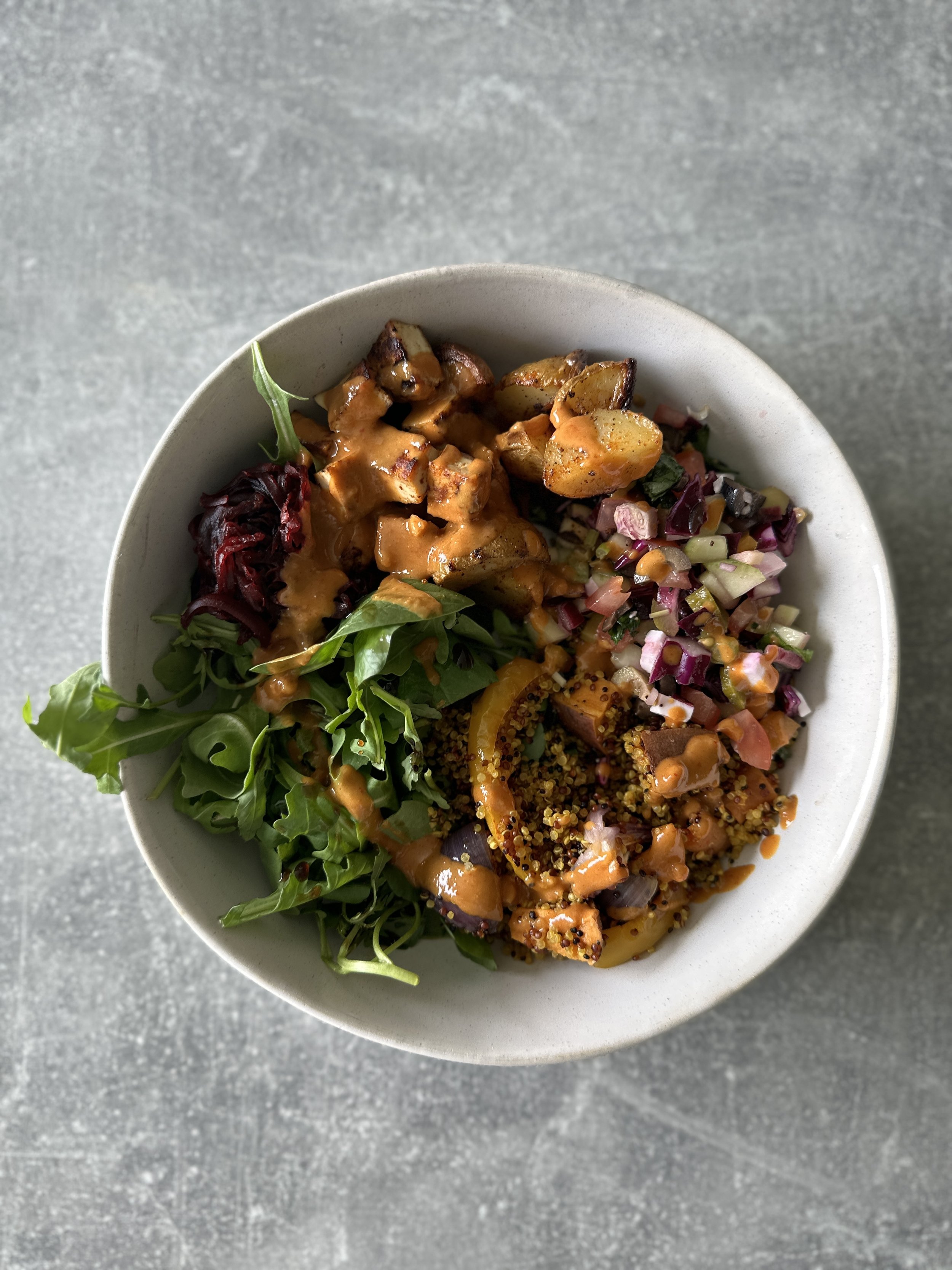 A bowl of mixed greens salad with quinoa, roasted vegetables, chopped onions, and a drizzle of dressing on top.