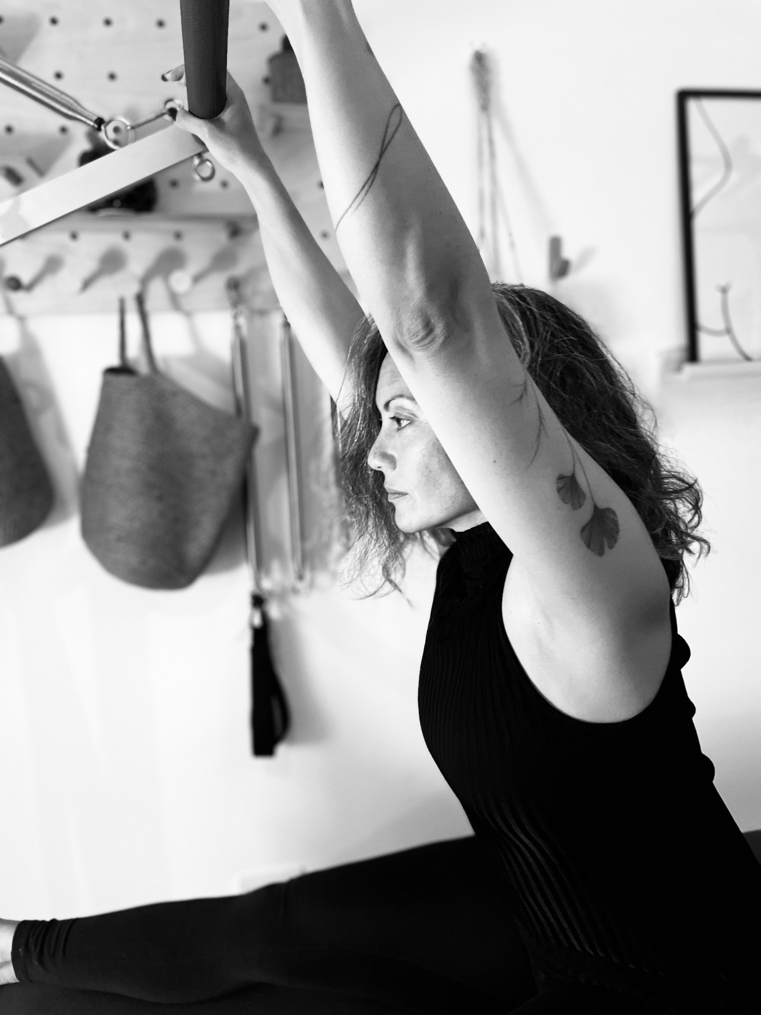A woman with curly hair sitting on the floor and stretching her arms overhead. She is in a workout or yoga pose in a home gym setting with workout equipment on the wall behind her.