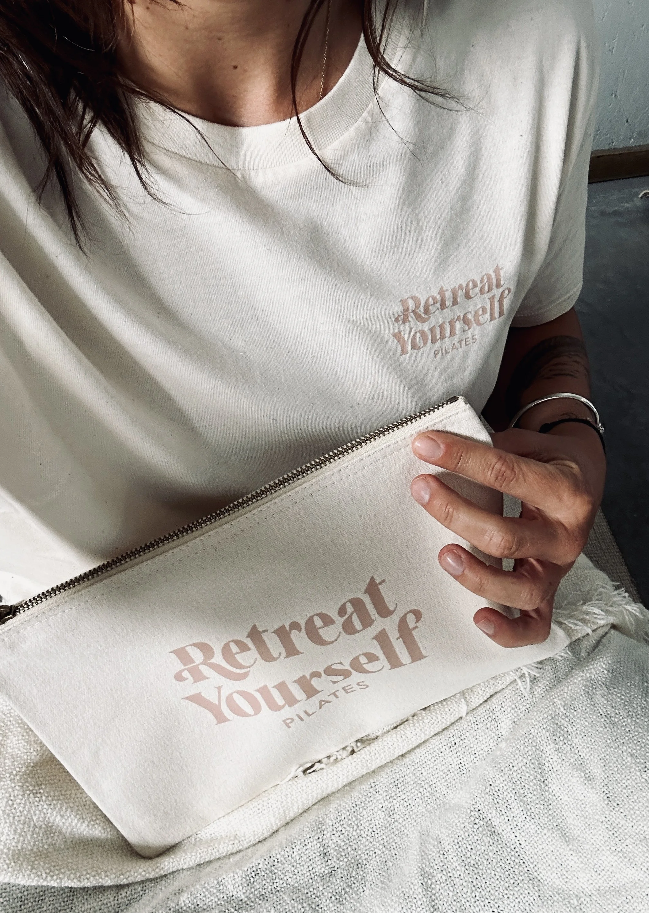 A person wearing a white t-shirt with the words "Retreat Yourself PILATES" and holding a matching cloth pouch with the same text, sitting on a light-colored surface.