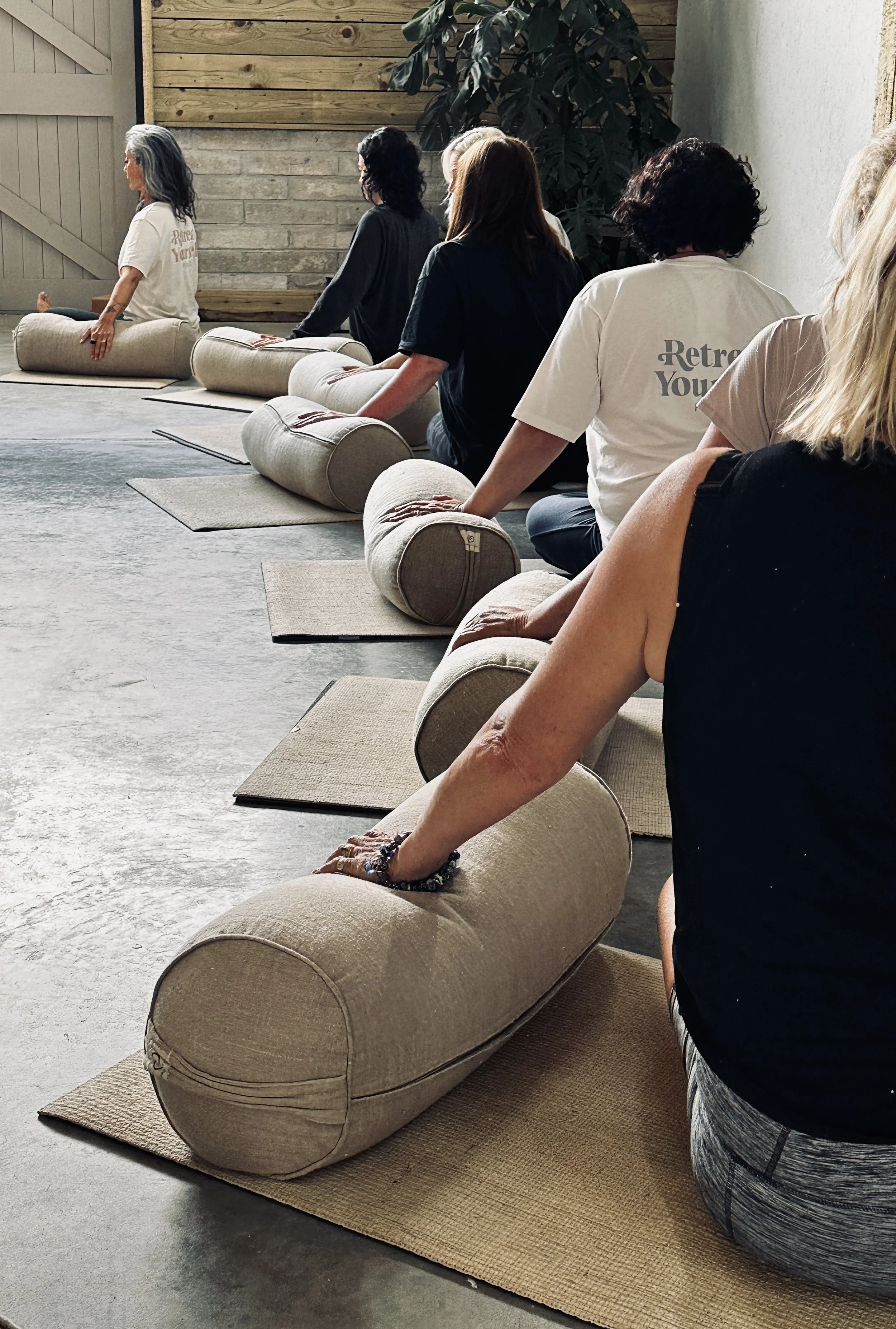 A group of people sitting on yoga mats with bolsters, participating in a meditation session in a studio with wooden walls and indoor plants.