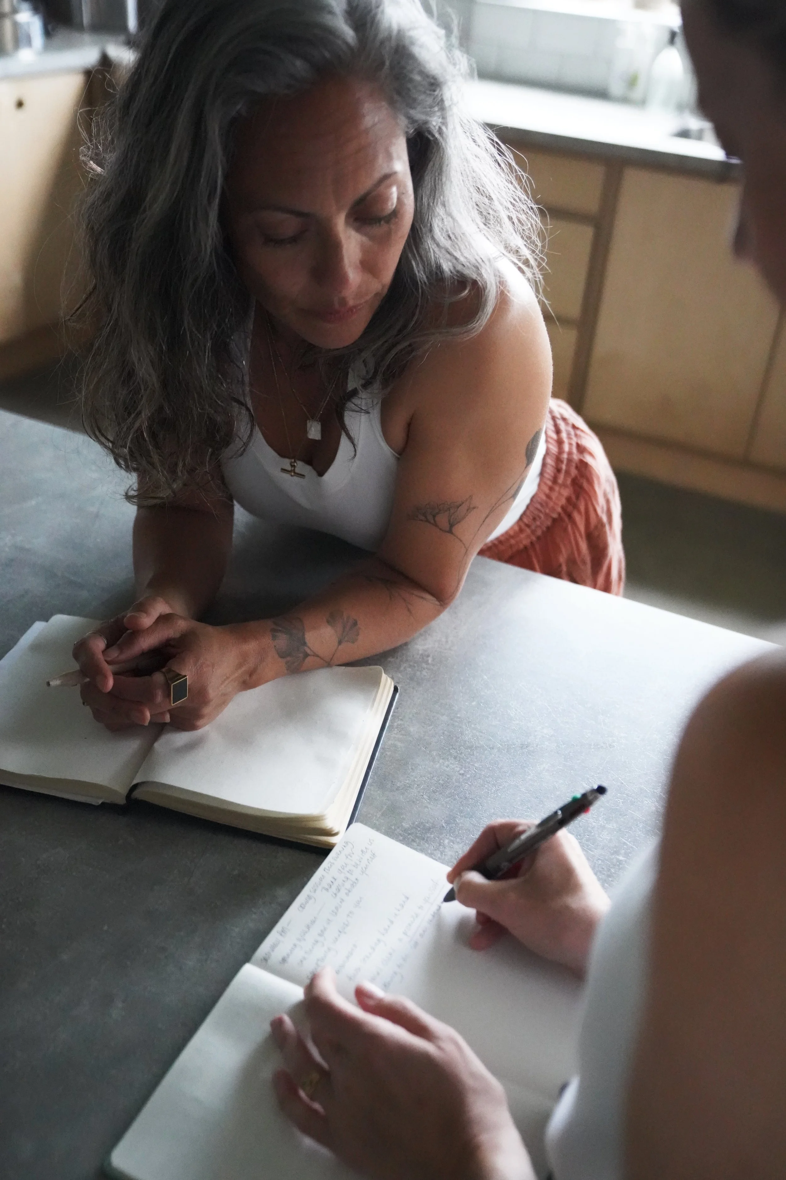 A woman with gray hair and tattoos leans over a table, writing in a notebook while talking to another person who is also writing in a notebook.