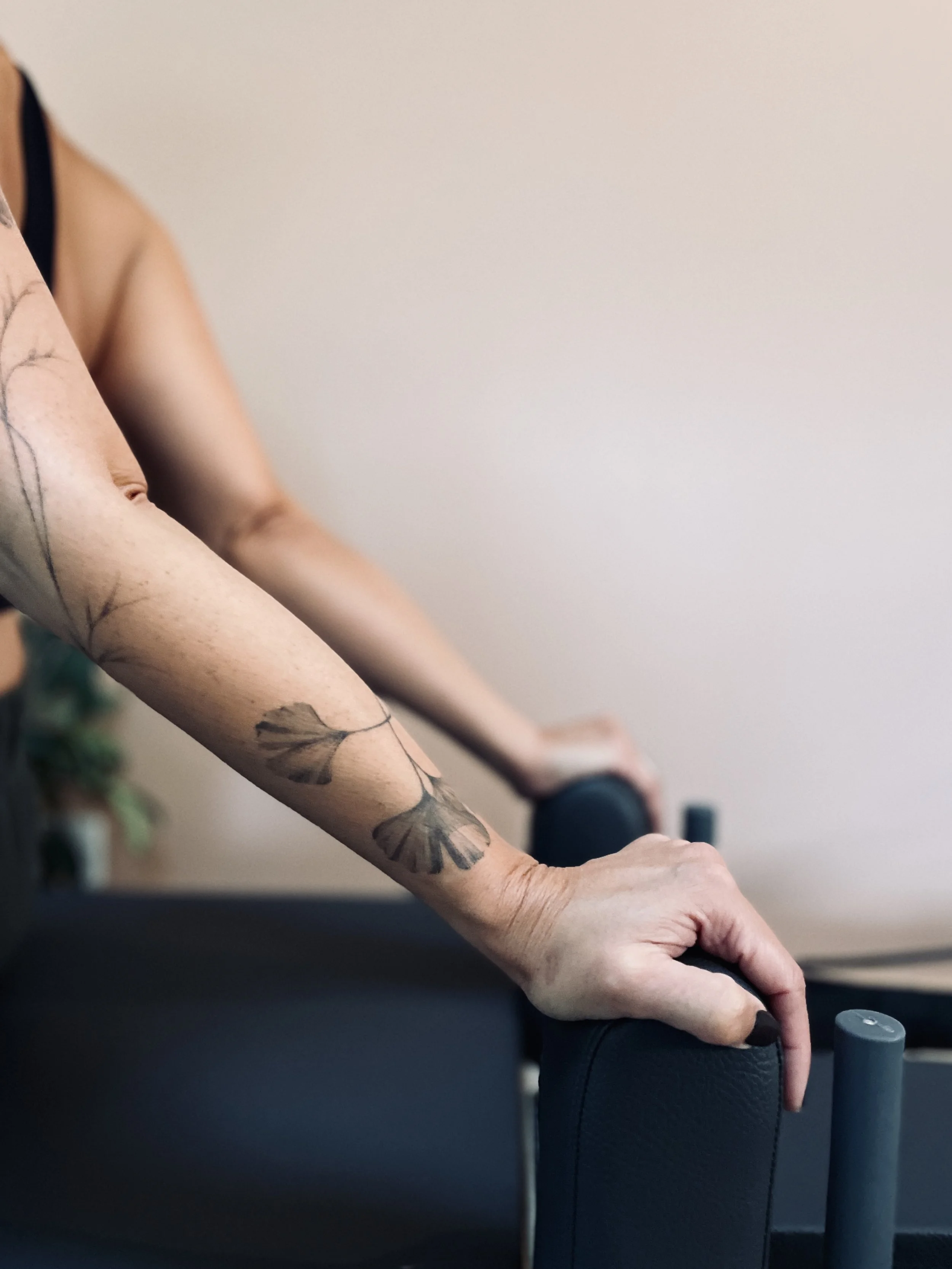 Close-up of a person's arm with a floral tattoo, holding the side of a black exercise or therapy chair, with a beige wall in the background.