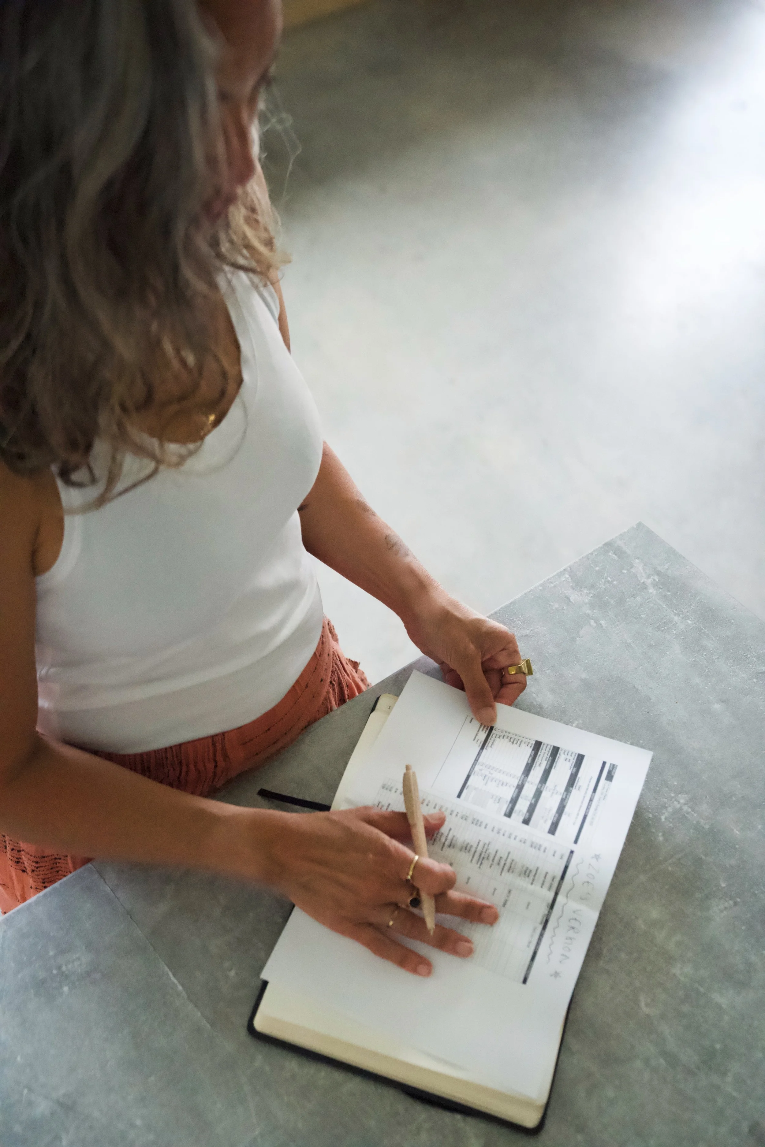 A woman standing at a table, writing in a notebook, with printed documents and a pen.