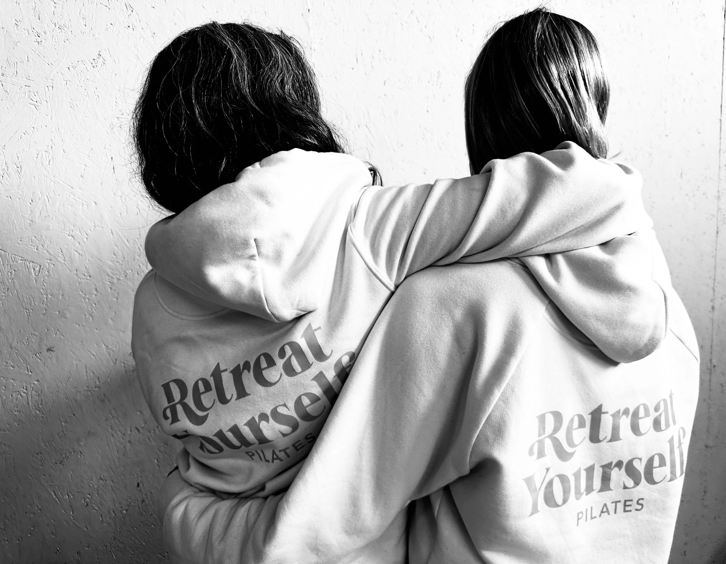 Two women wearing hoodies with the words "Retreat Yourself Pilates" printed on the back, standing close together, facing away from the camera, against a textured wall.