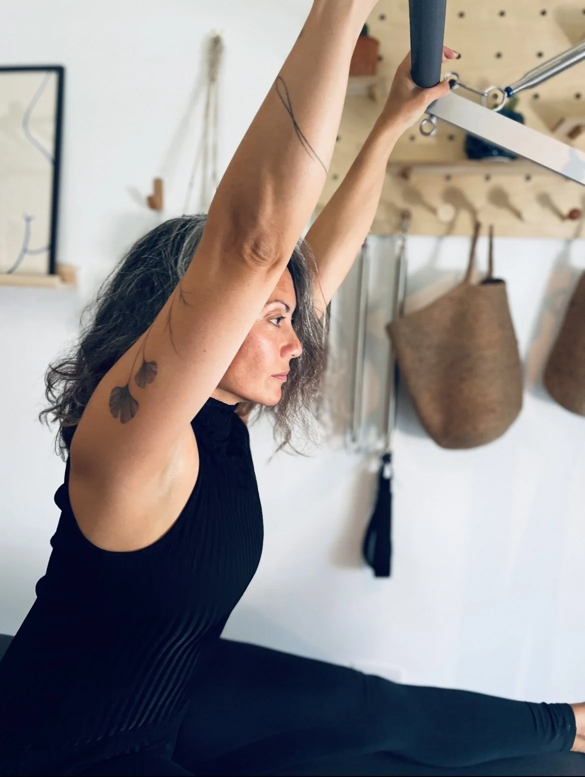 Woman with curly hair in a black tank top performing a stretch in a studio with wall-mounted storage baskets and hanging tools.