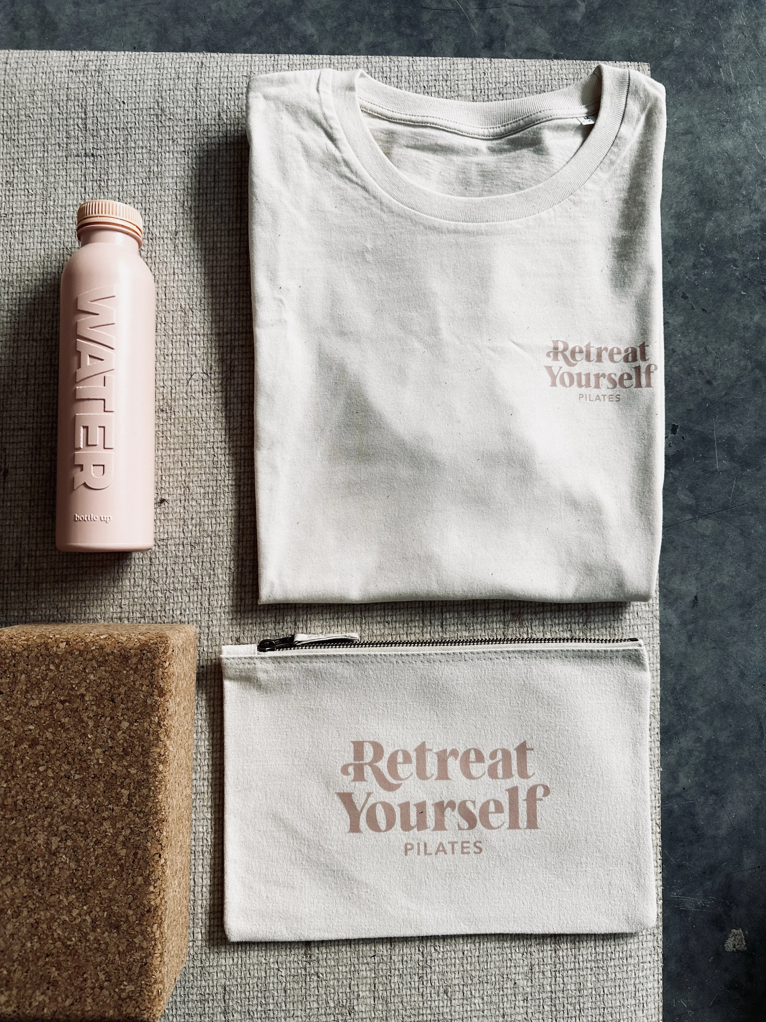 A flat lay of a white t-shirt, a pink water bottle, a white zippered pouch, and a brown cork yoga block on a textured surface. The t-shirt and pouch both have the logo 'Retreat Yourself Pilates' printed on them.