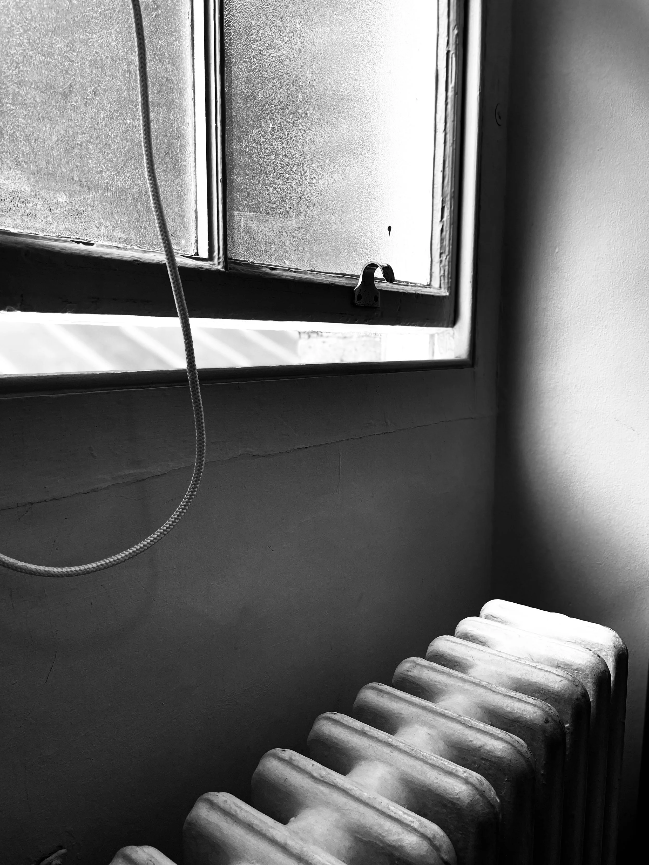 A black and white photo of an open window with a latch, next to a wall and an old radiator. The window is slightly fogged or dirty, with some sunlight coming through. A hanging cord is visible on the left side of the image.