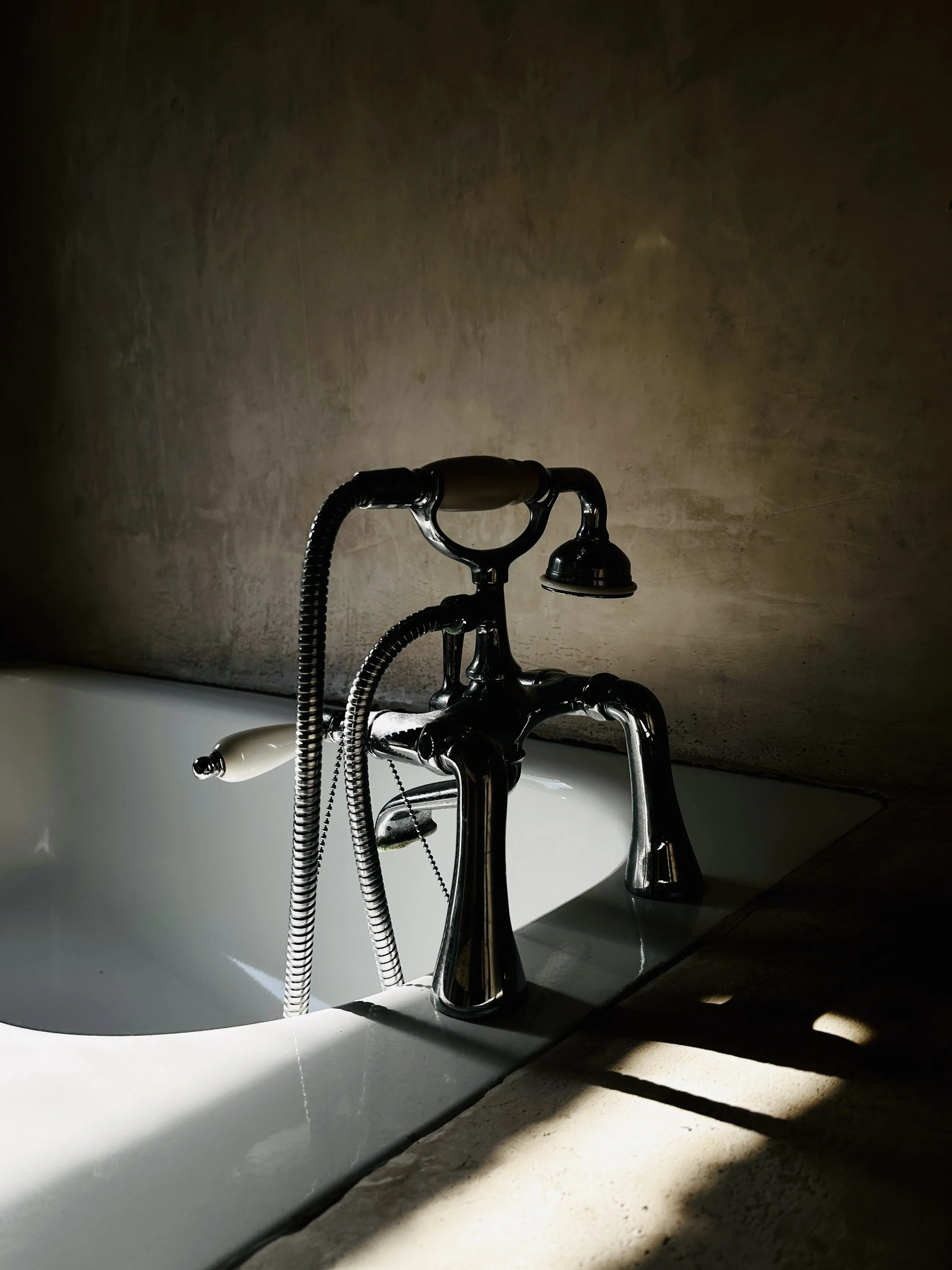 Close-up of a vintage-style bathtub faucet with a handheld showerhead, set against a textured wall and illuminated by natural light, creating shadows on the tub's edge.