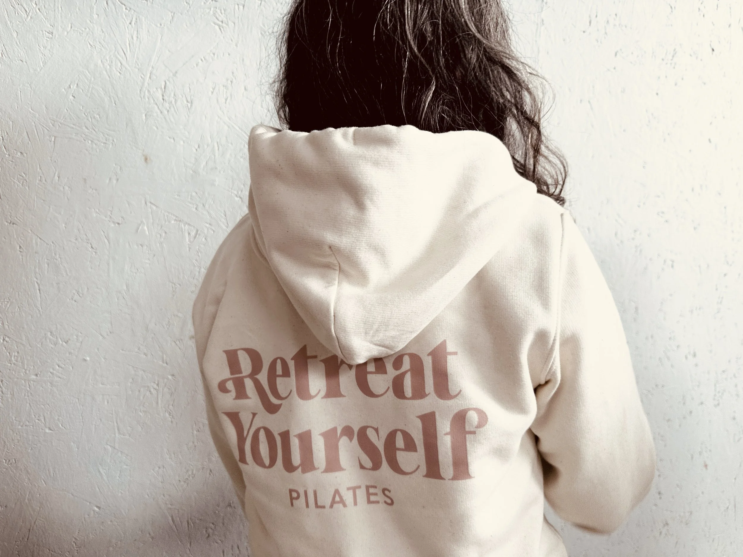 Back of a person wearing a cream-colored hoodie with the words 'Retreat Yourself Pilates' printed on it, standing against a textured white wall.