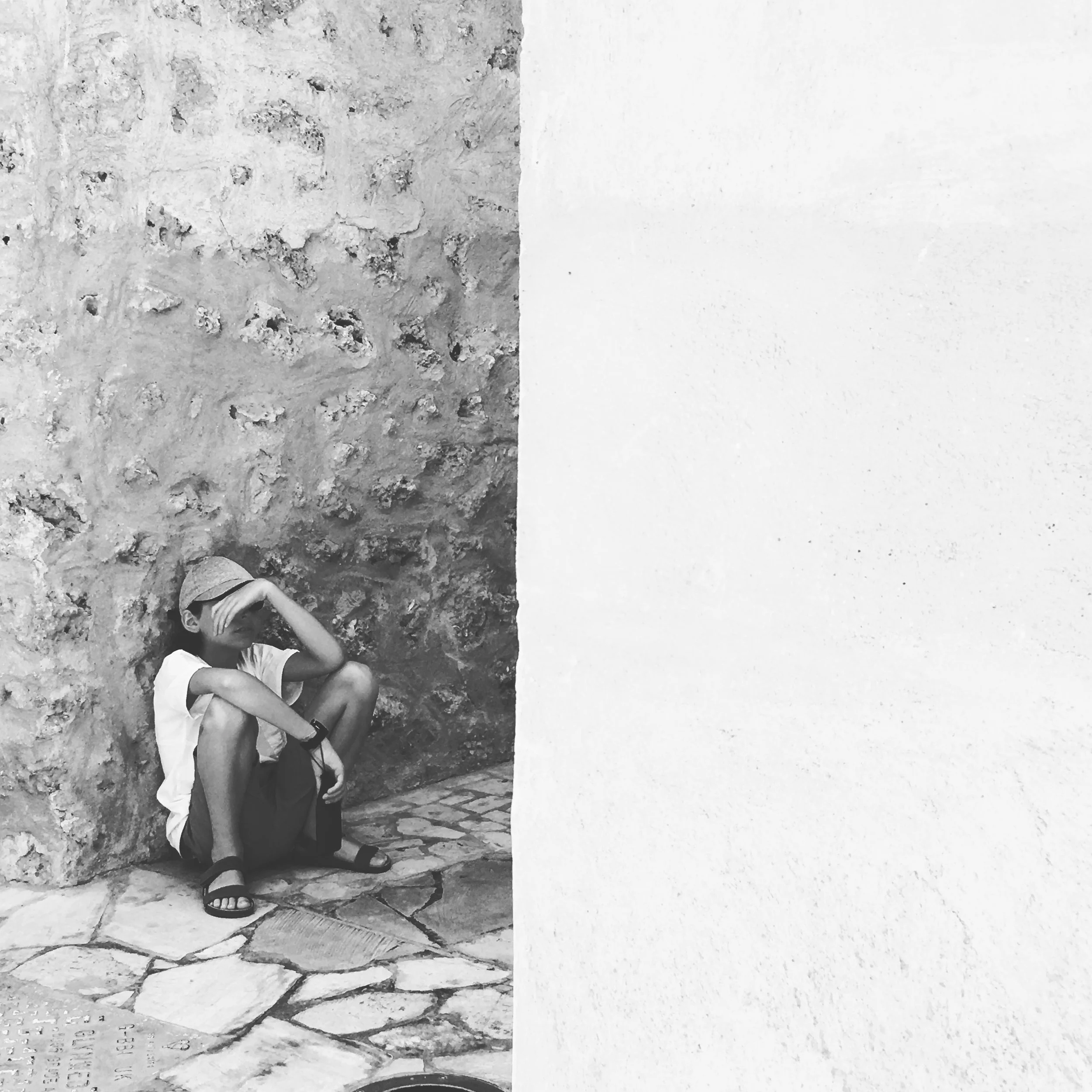 A boy sitting on a stone pavement, leaning against a textured stone wall, with one arm resting on his knee and his hand covering his face.