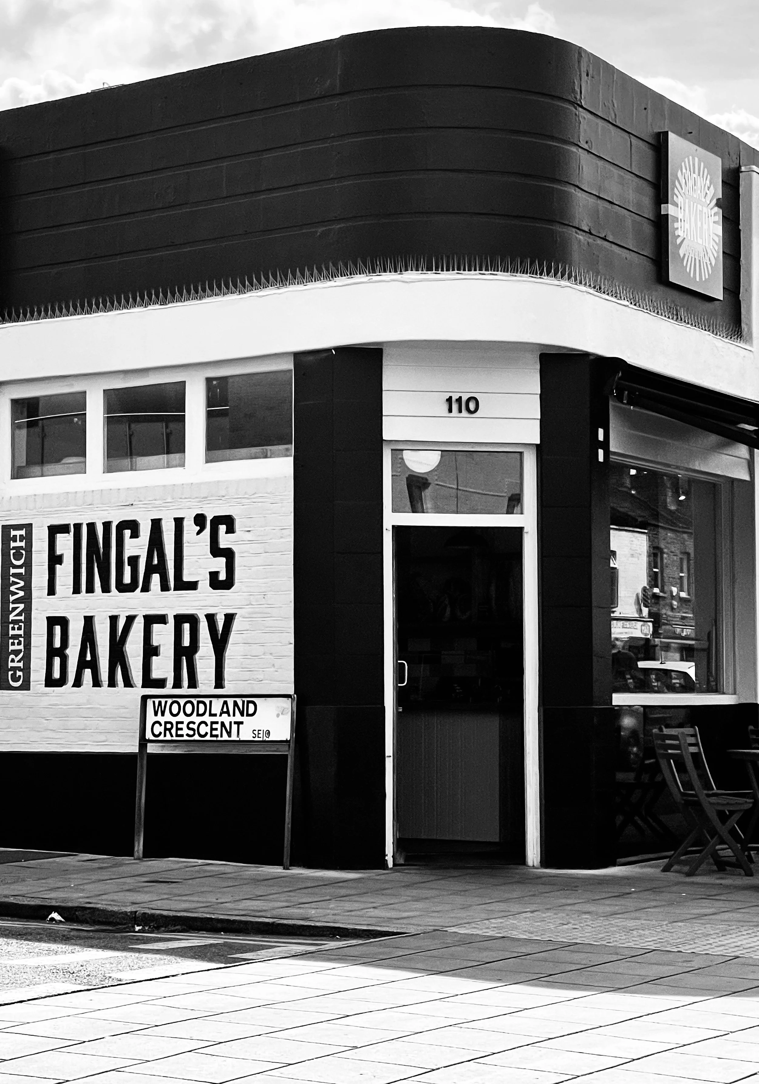 Black and white photo of Fingal's Bakery, located at 110 Greenwood Street, with a sign indicating the street name Woodland Crescent. The bakery has large windows, a door in the middle, and outdoor tables with chairs.