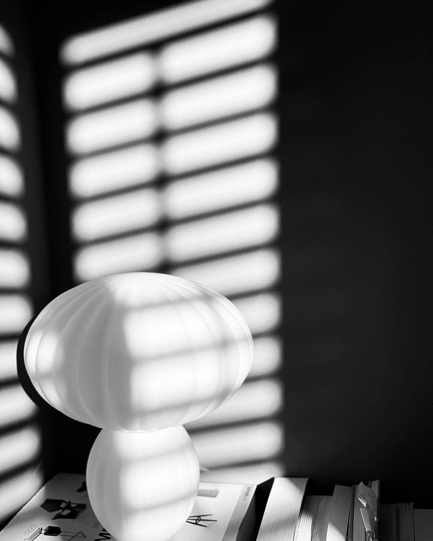 A modern lamp with a layered design placed on top of magazines or books, casting shadows through window blinds on a dark wall.