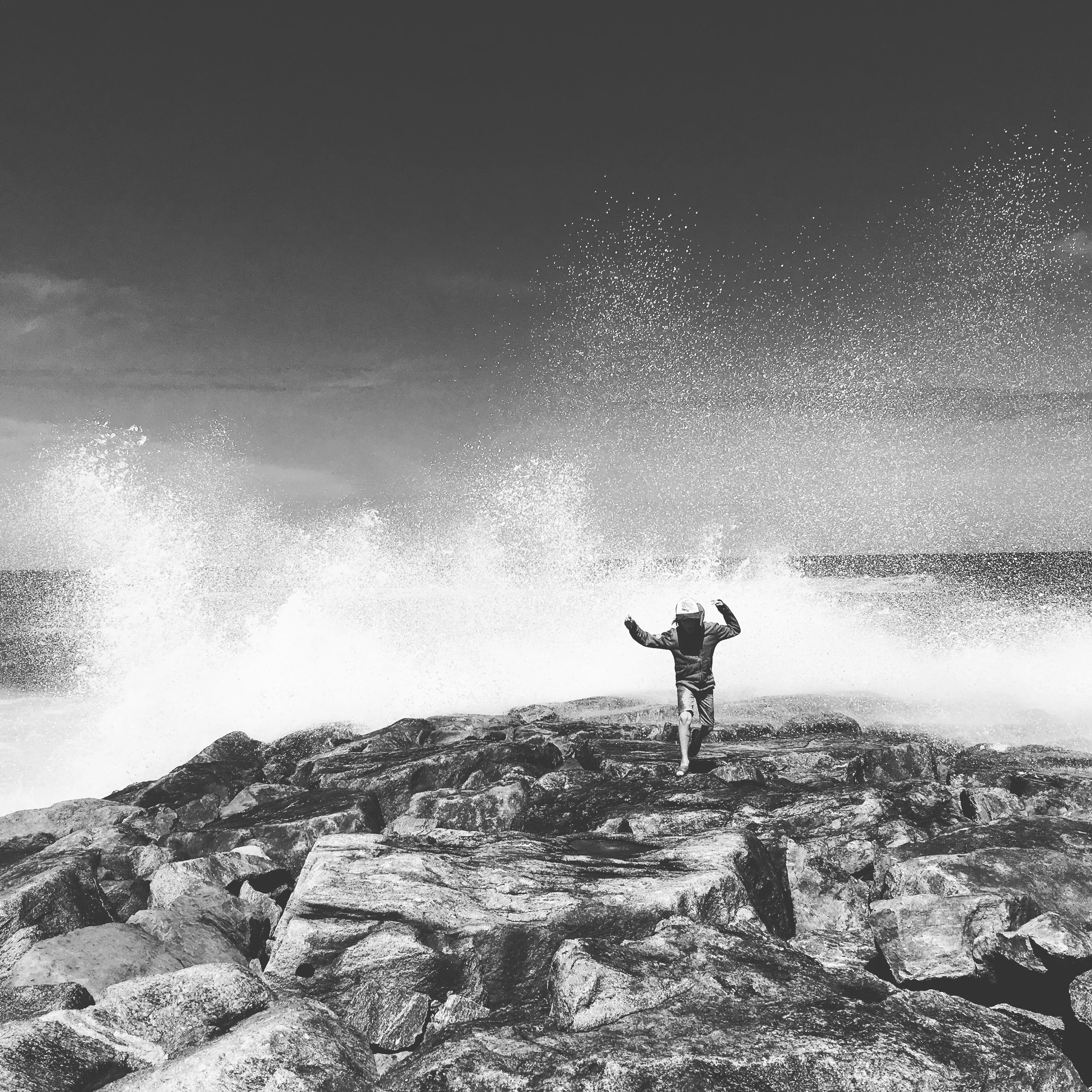 A person running on rocks by the ocean, with waves crashing behind them and spray in the air, under a cloudy sky.