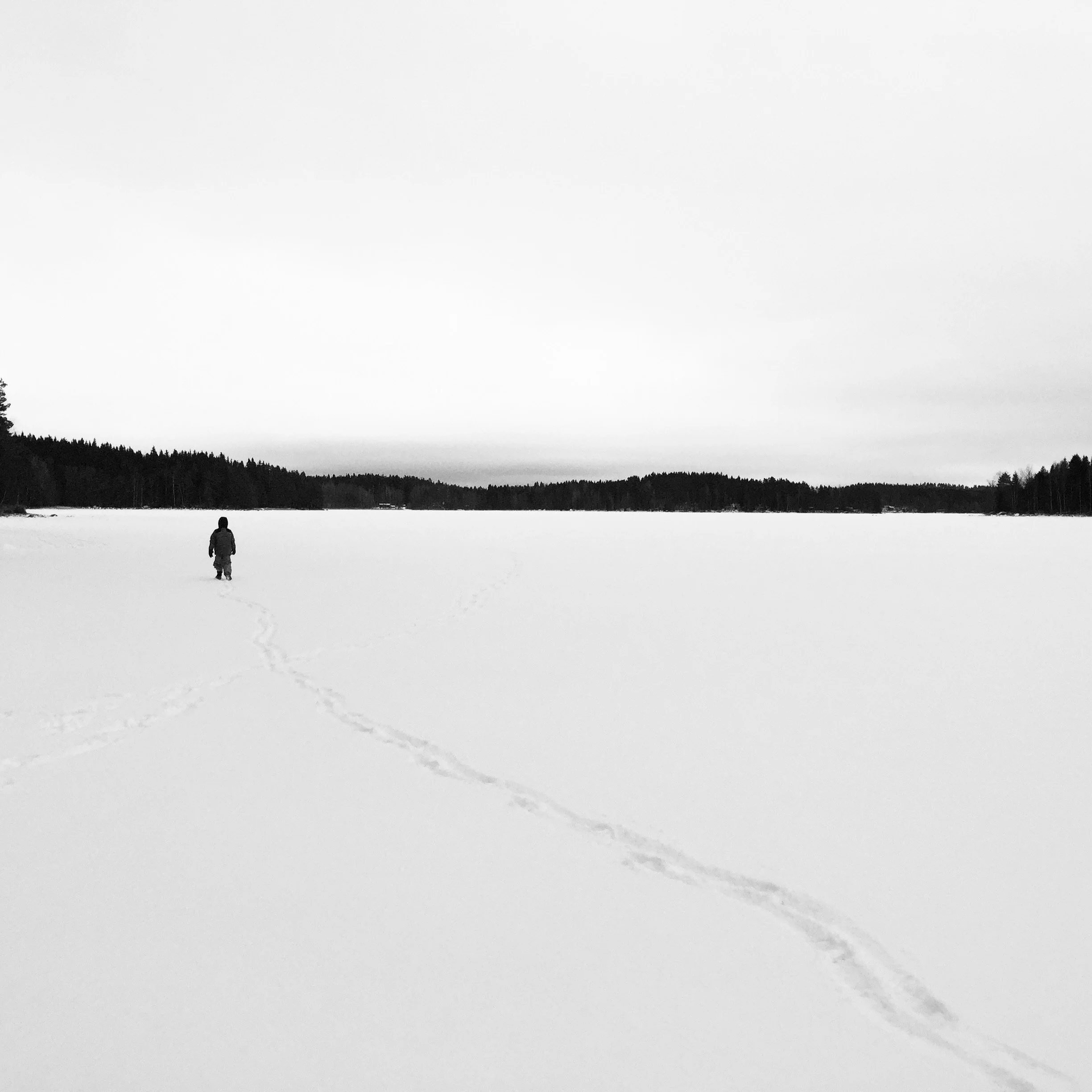 A person walking alone across a snow-covered lake under an overcast sky, with tracks visible in the snow, and a forested shoreline in the distance.