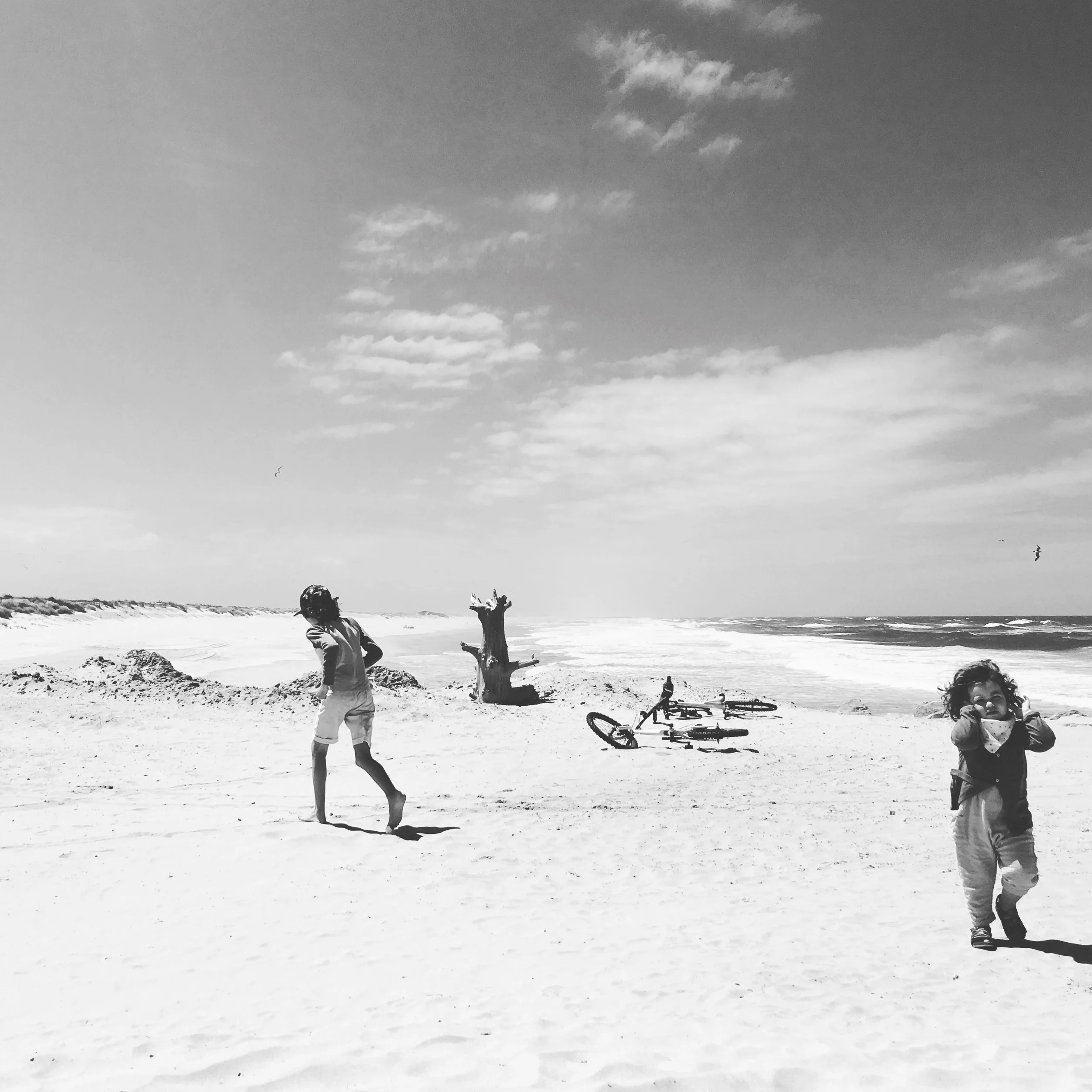 Black and white photo of children playing on a sandy beach with bikes and driftwood, ocean waves in the background, and a sky with scattered clouds.