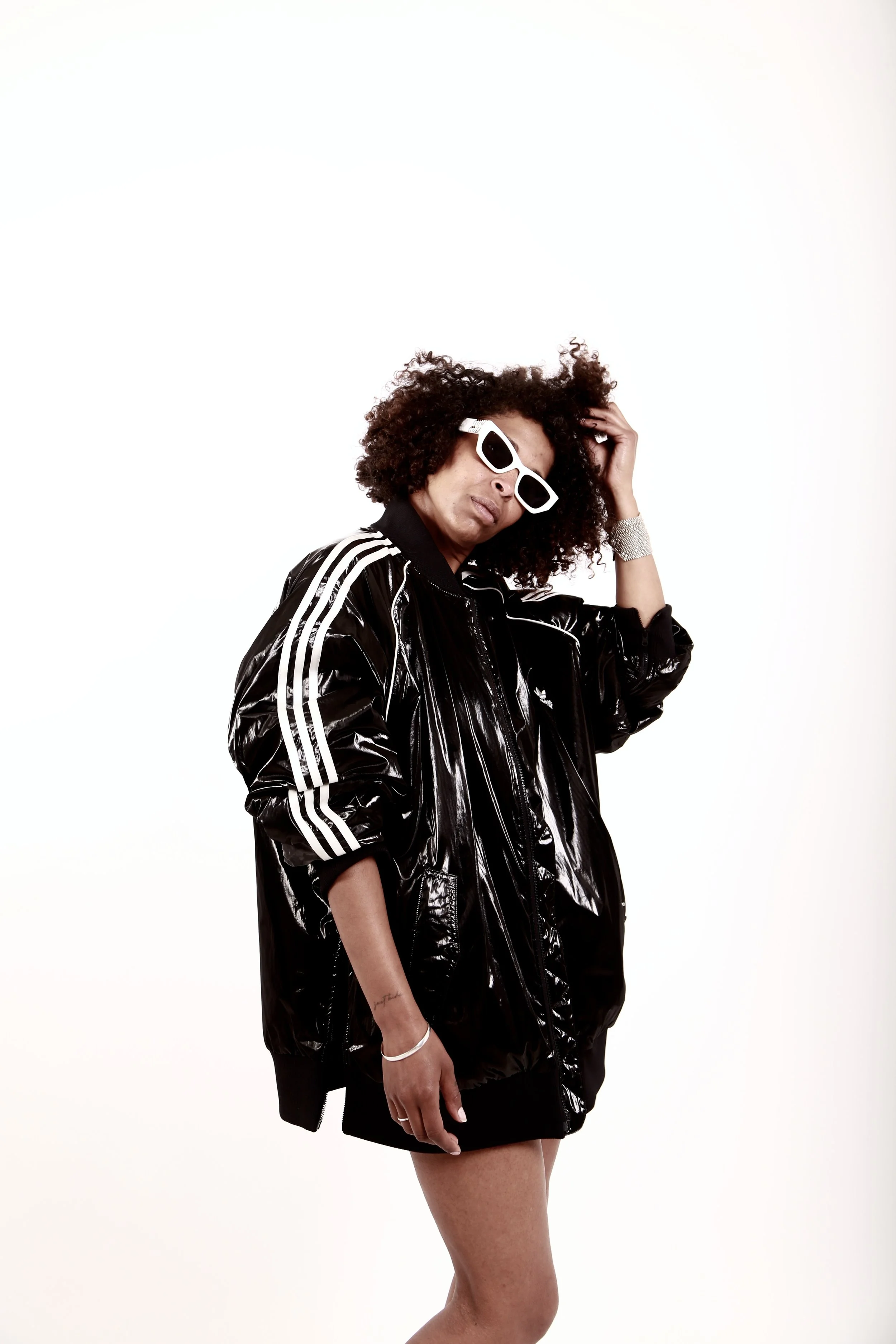 Person wearing a black shiny jacket, white sunglasses, and a silver bracelet, standing against a white background.