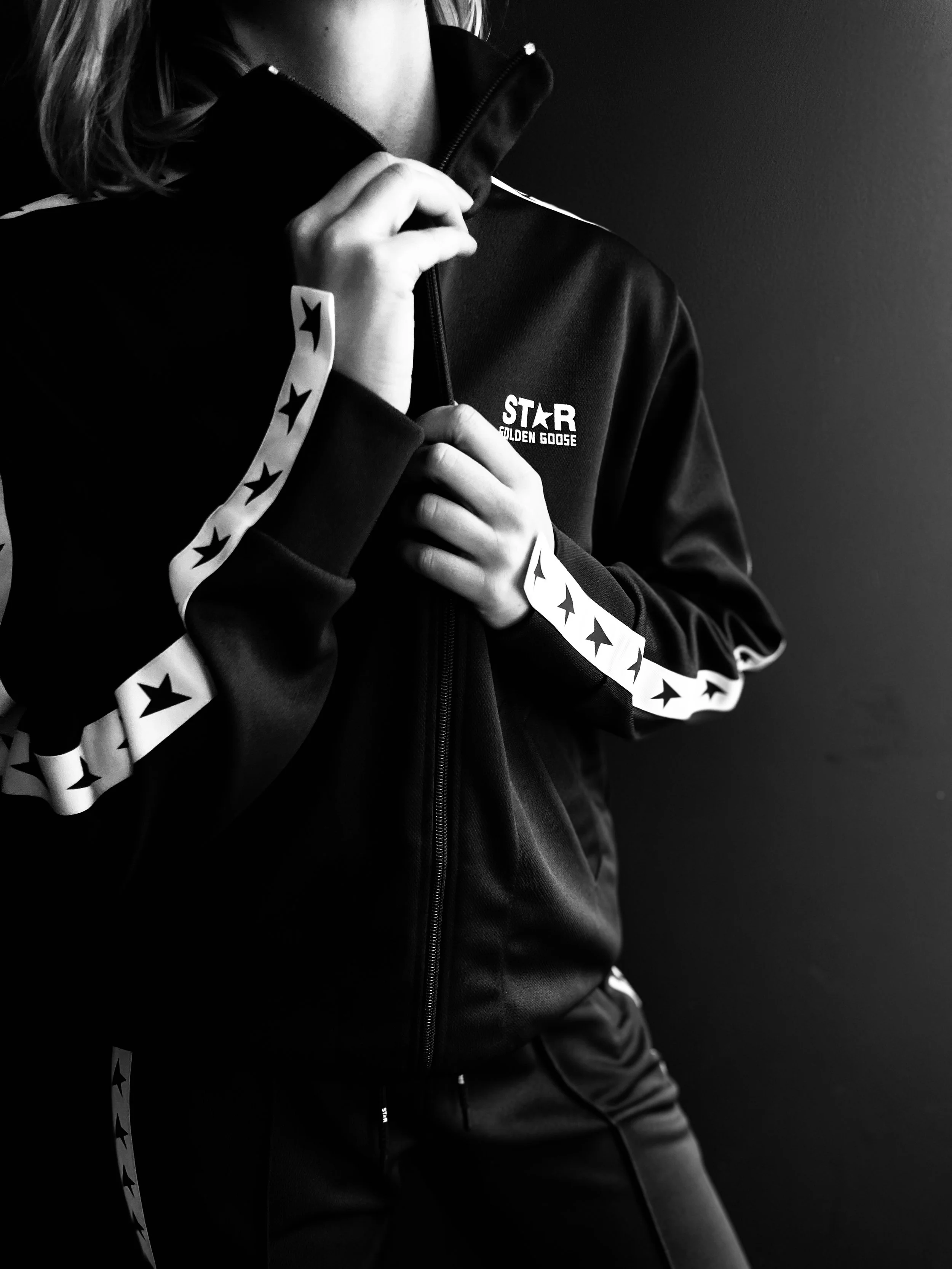Close-up of a woman wearing a black jacket with white star patterns on the sleeves, zipping up the jacket against a dark wall.
