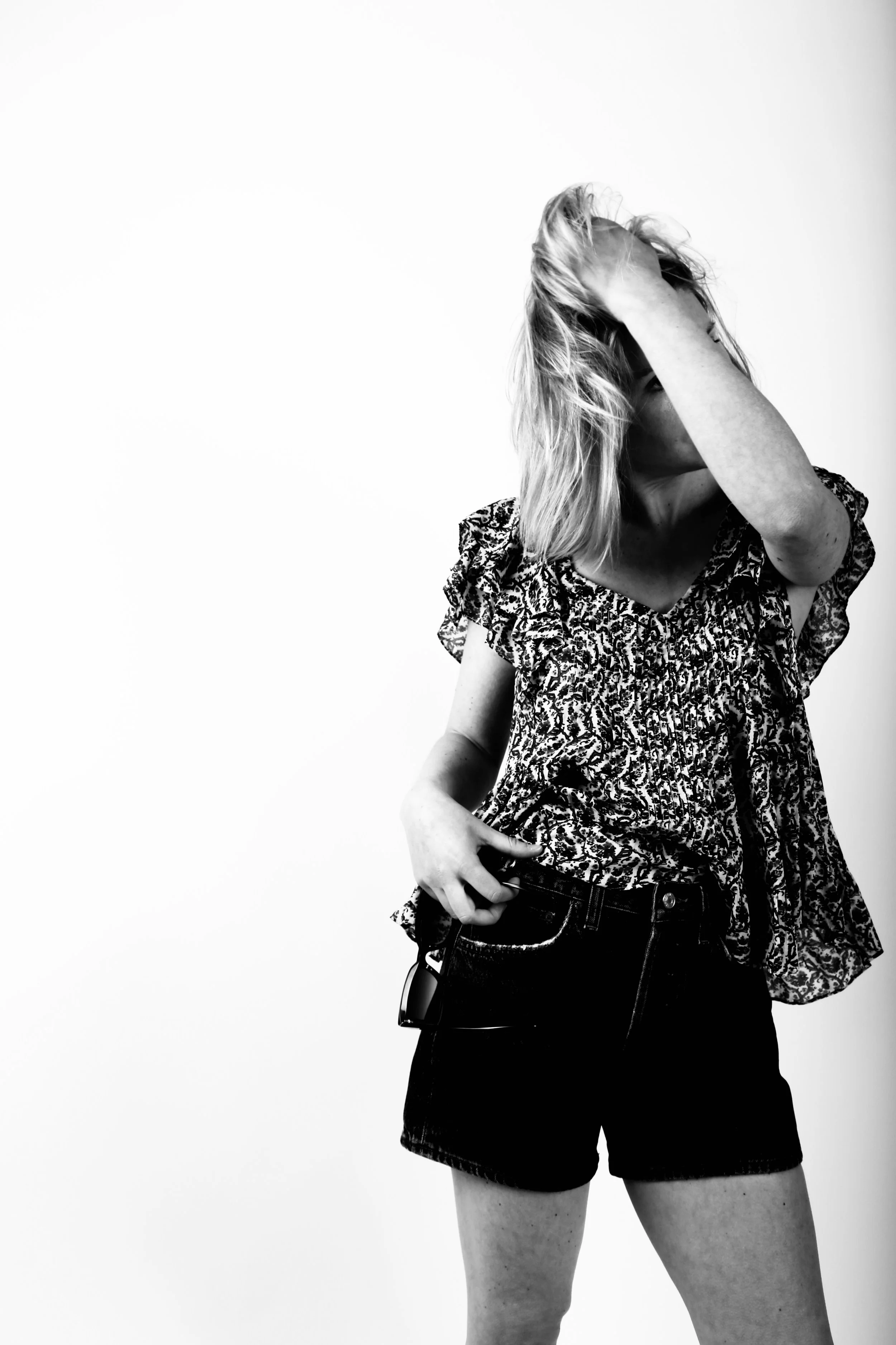 A woman in a patterned shirt and black shorts standing against a plain white background, partially covering her face with her hair.