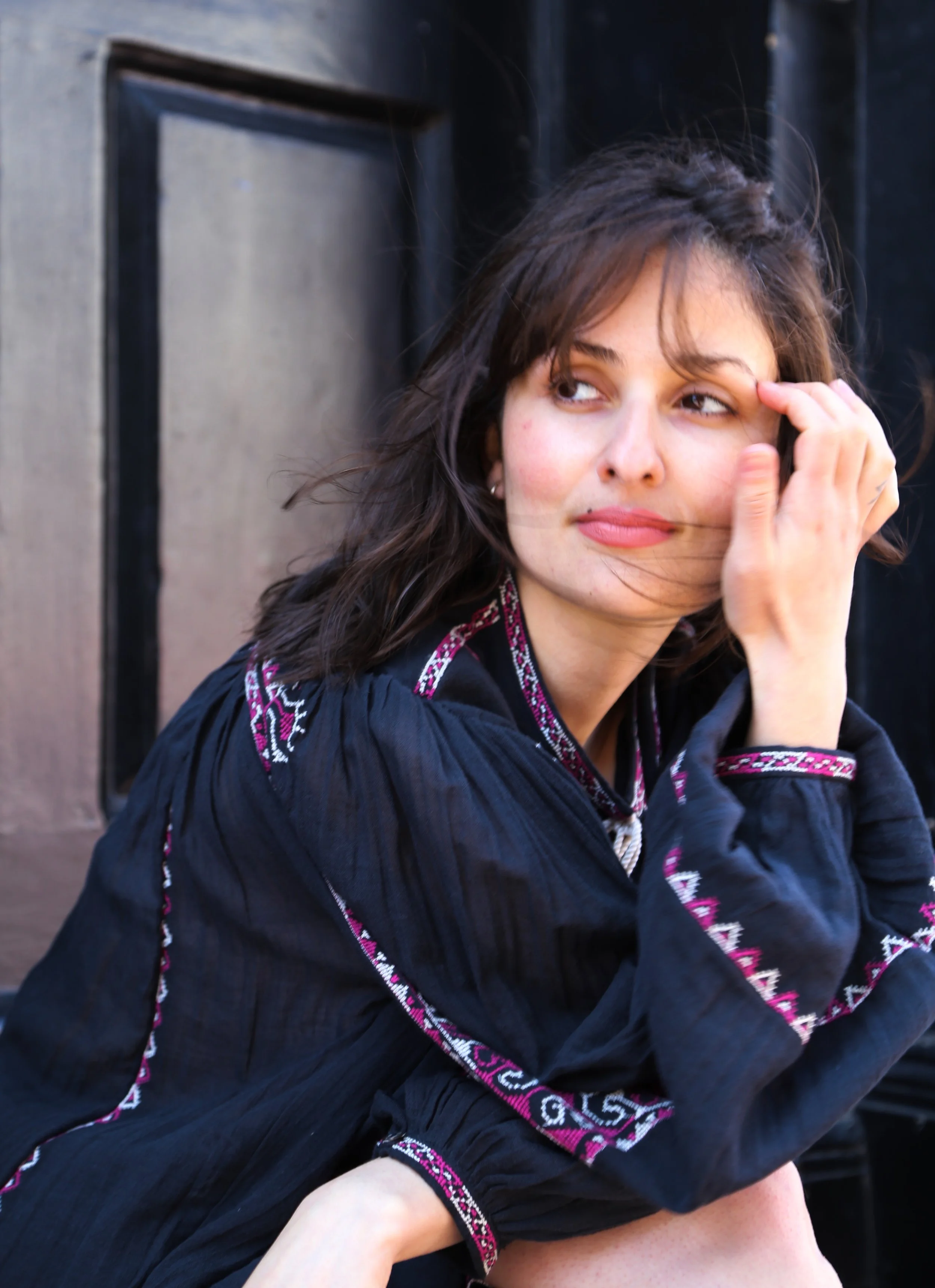 A woman with brown hair and light skin sitting outdoors near a dark door, wearing a black embroidered dress, touching her face and looking to her left.