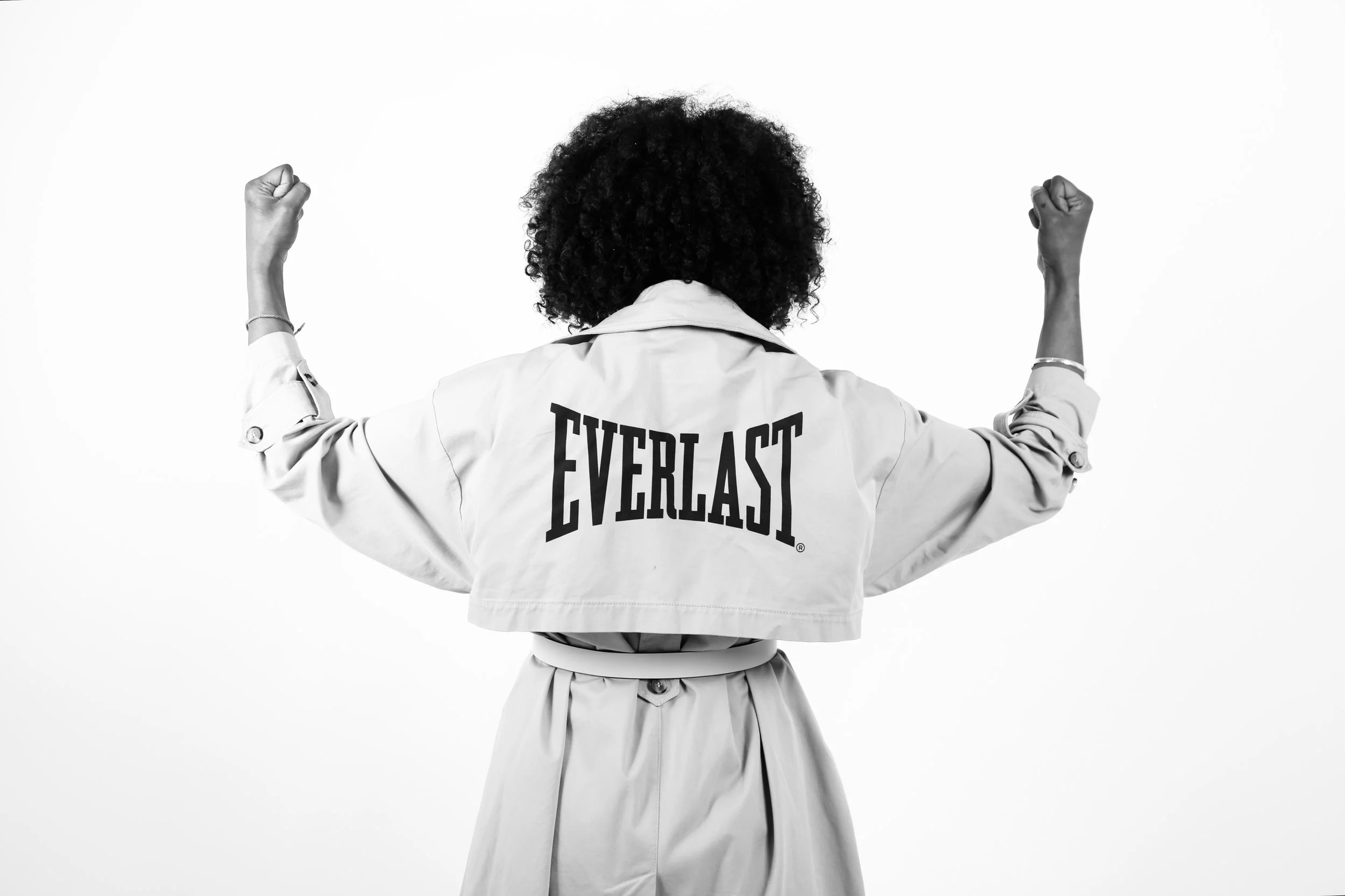 Back view of a woman with curly hair flexing her arms, wearing a jacket with the 'Everlast' logo on the back.