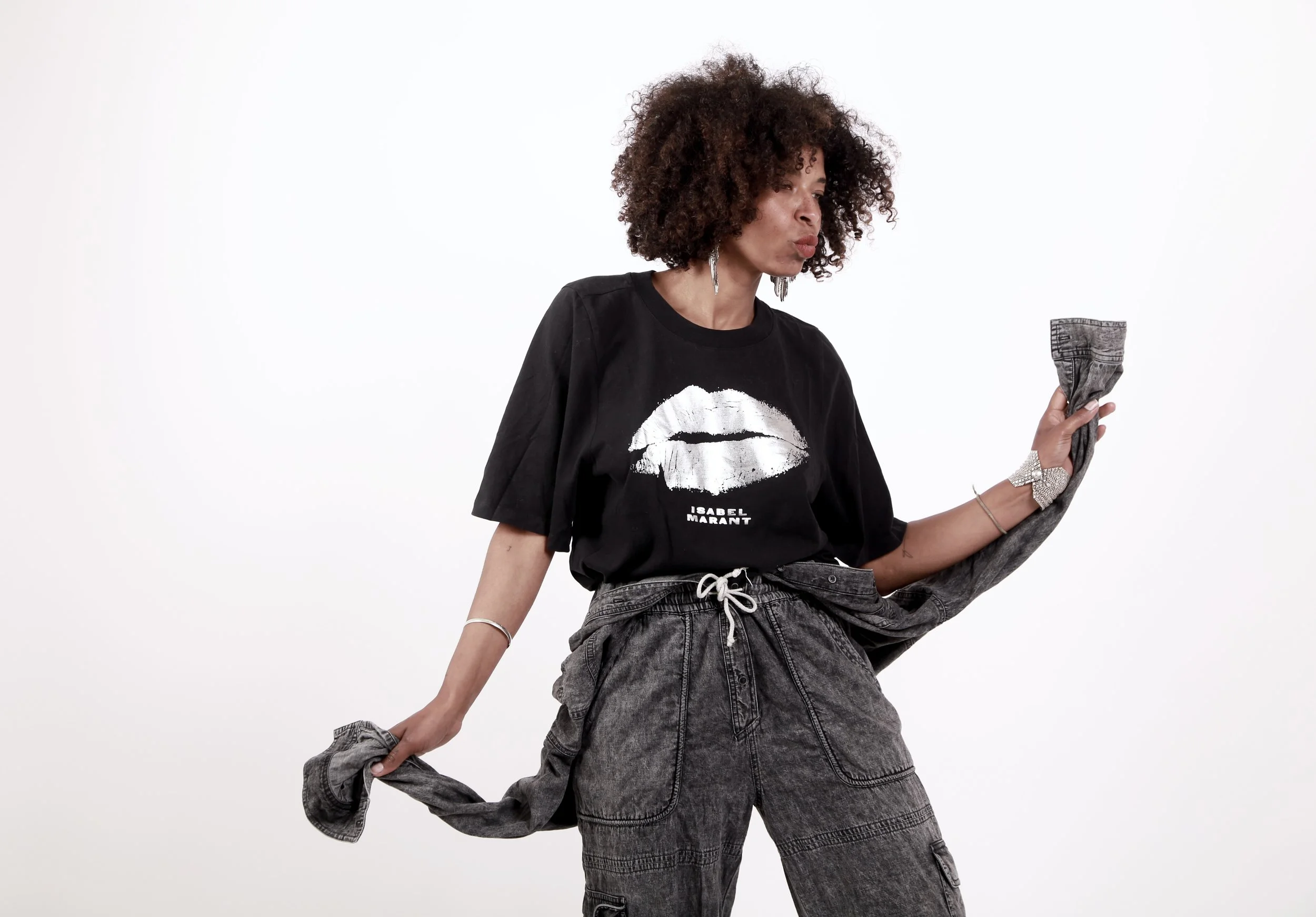 A woman with curly brown hair wearing a black t-shirt with white lips print and the words 'Isabel Marant,' and gray baggy cargo pants, holding a rolled-up denim jacket in a studio against a white background.
