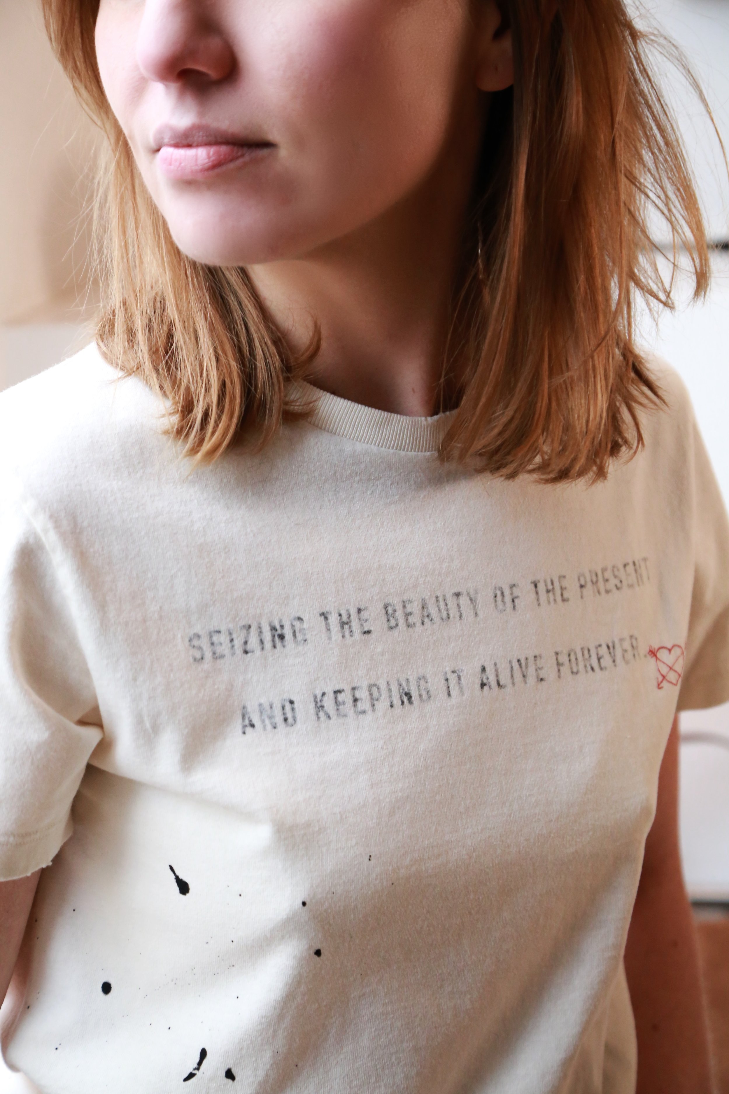 Close-up of a woman's chest and face, wearing a cream-colored T-shirt with text that reads 'Seizing the beauty of the present and keeping it alive forever.'