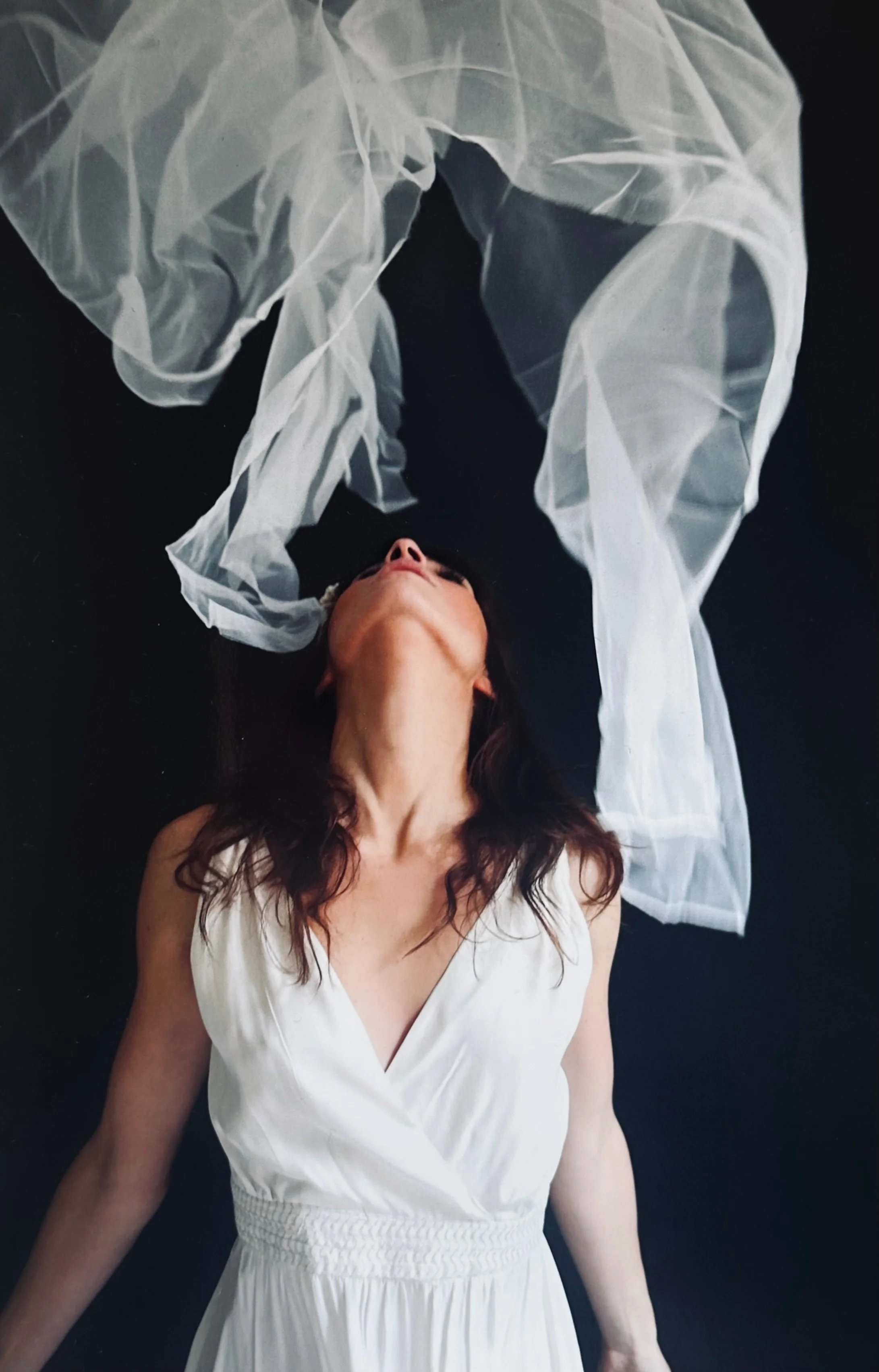 A woman with dark hair and light skin lying on a black surface with her head tilted back, wearing a sleeveless white dress. A sheer, flowing white fabric is draped above her, creating an abstract, artistic scene.