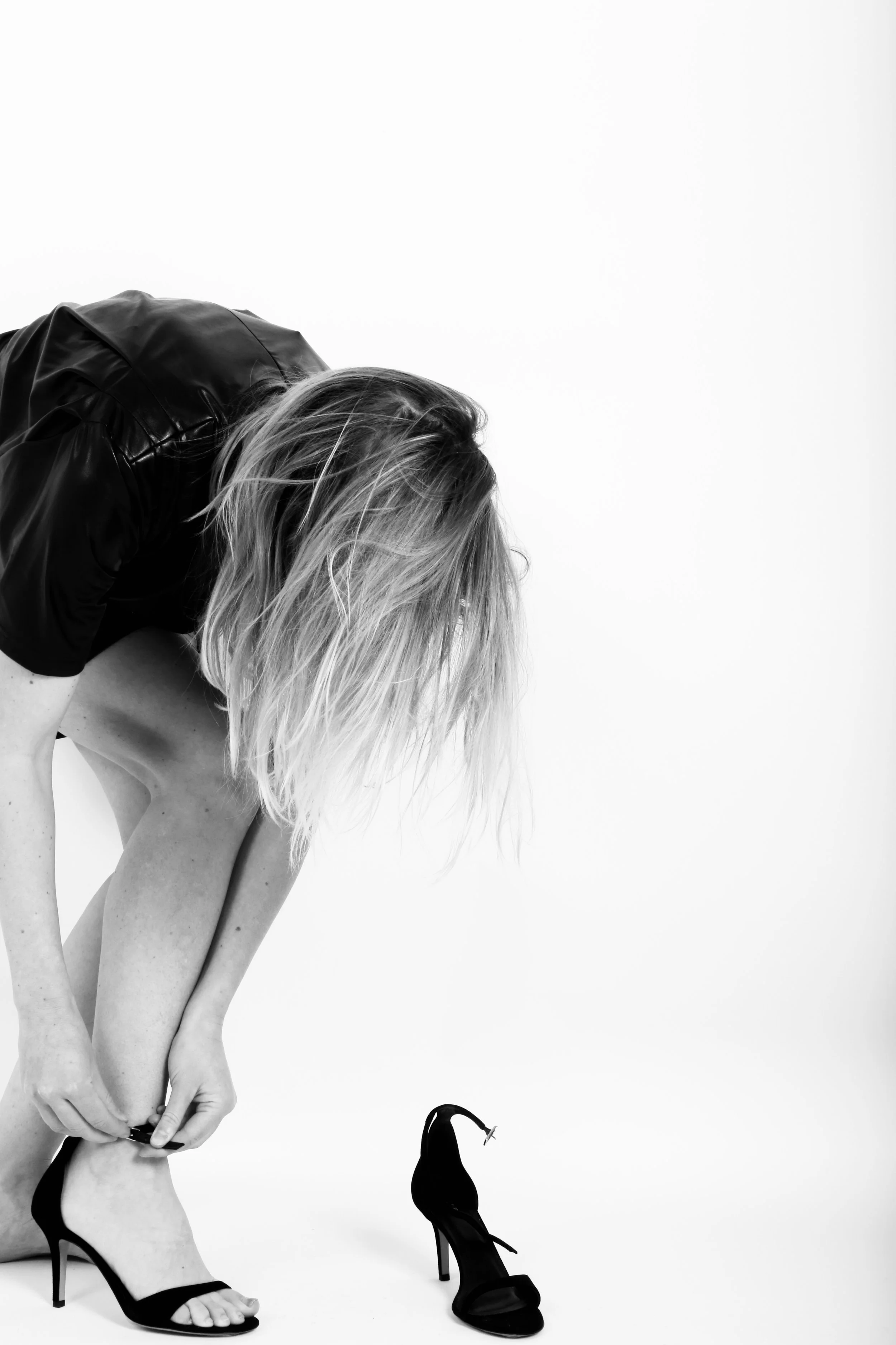 Black and white photo of a woman with blonde hair, wearing a black leather jacket, bending down to put on a high-heeled sandal. A second empty high-heeled sandal is on the floor nearby against a plain white background.