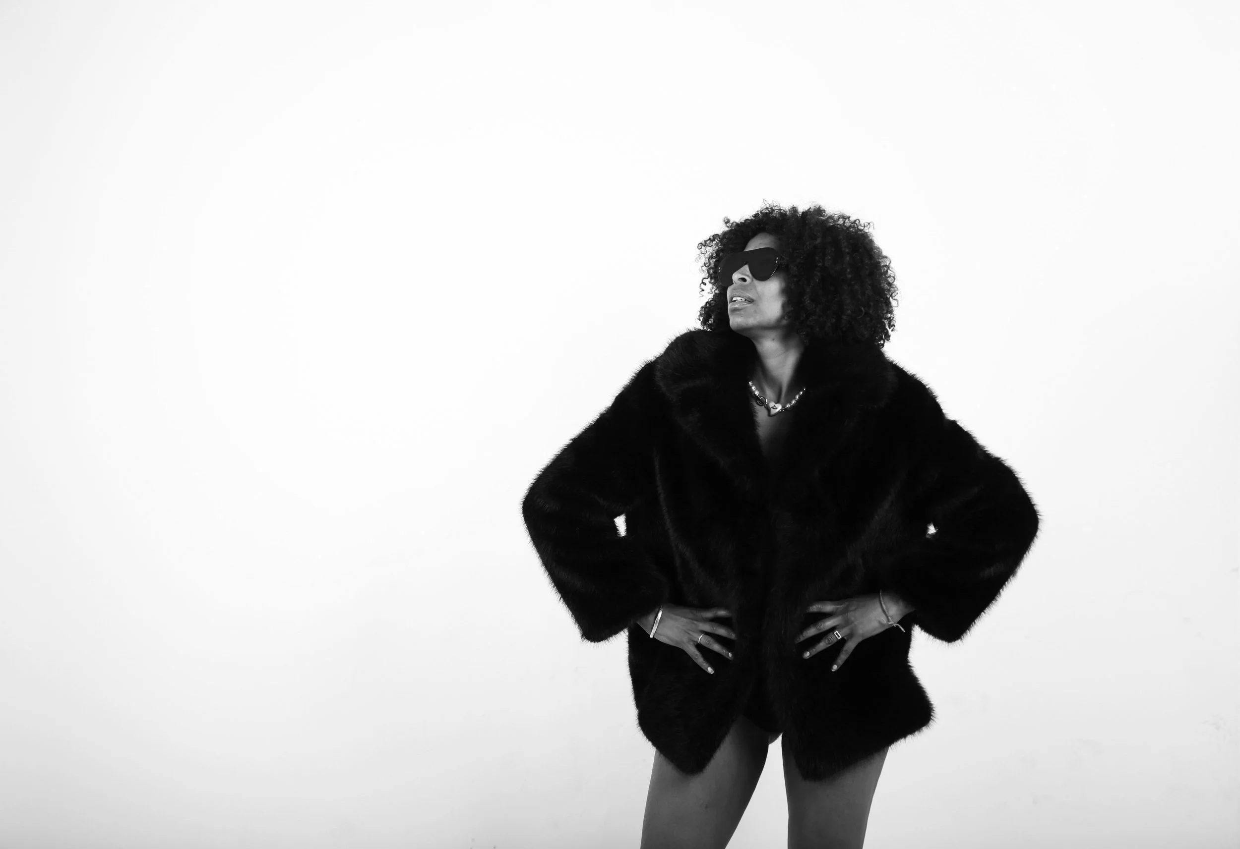 A woman with curly hair wearing sunglasses, a fur coat, and jewelry, standing with hands on hips against a plain white background.