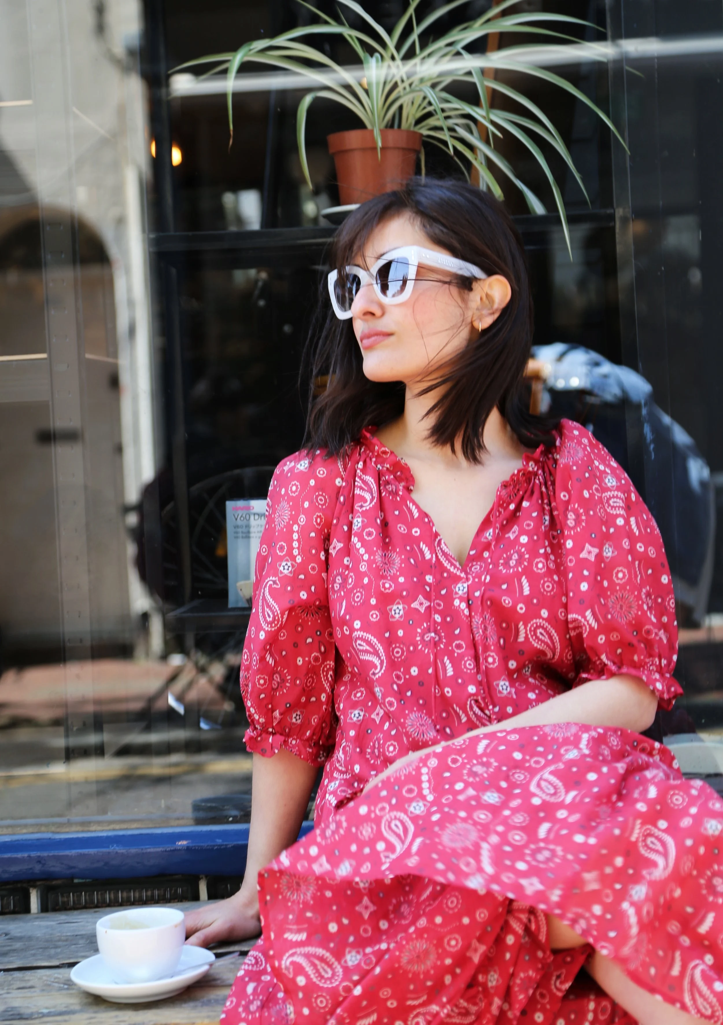 A woman in a red dress with white paisley patterns sitting outside, looking to the side, wearing white sunglasses, with a potted plant behind her and a tea cup on the table.