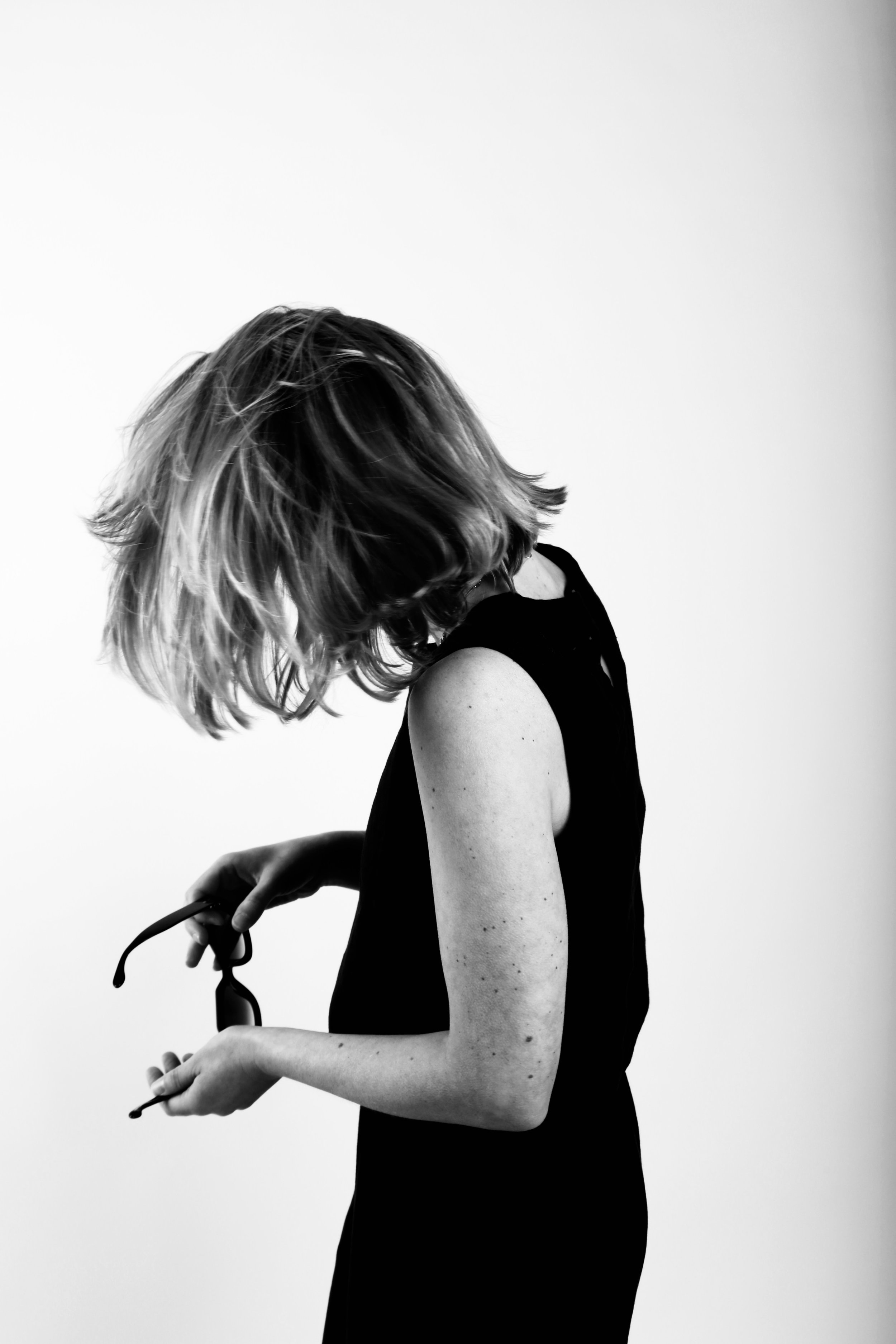 A woman with shoulder-length hair holding sunglasses in her hands, wearing a sleeveless black top, against a plain white background.