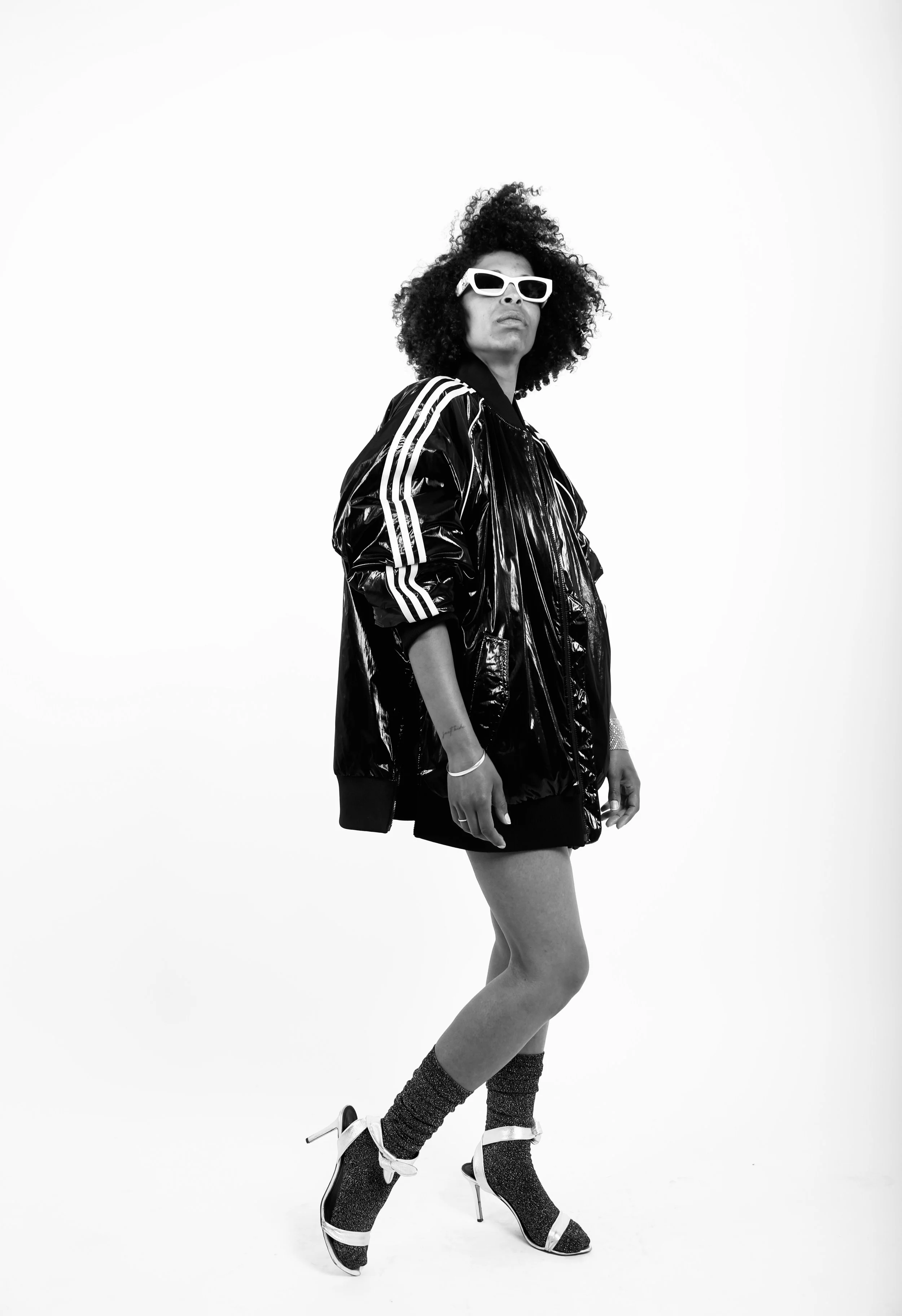 A woman with curly hair wearing white sunglasses, a shiny black jacket, black shorts, sparkly socks, and high heels, standing against a plain white background.