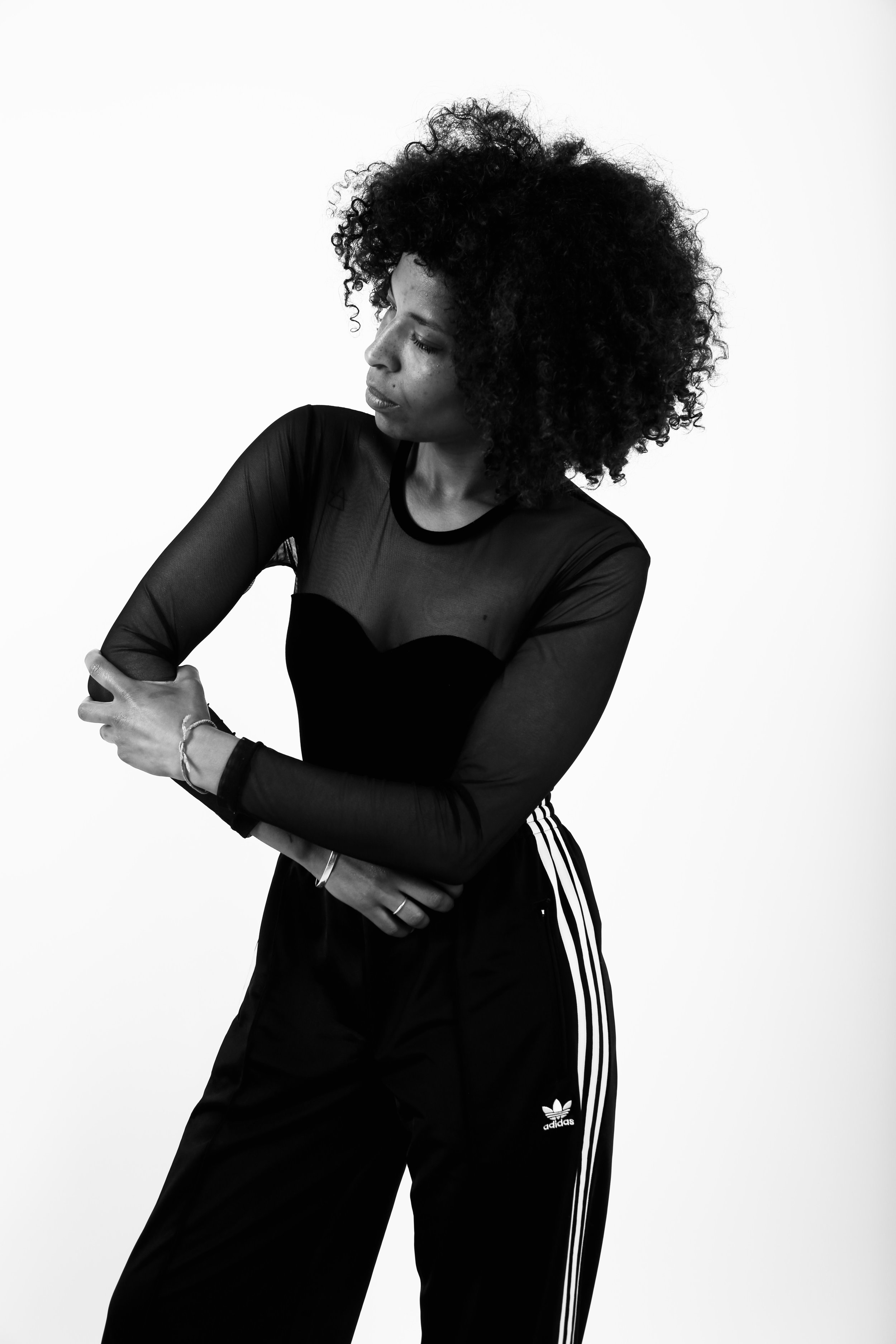 Black and white portrait of a woman with curly hair, wearing a sheer long-sleeve top and Adidas track pants, looking down and crossing her arms.