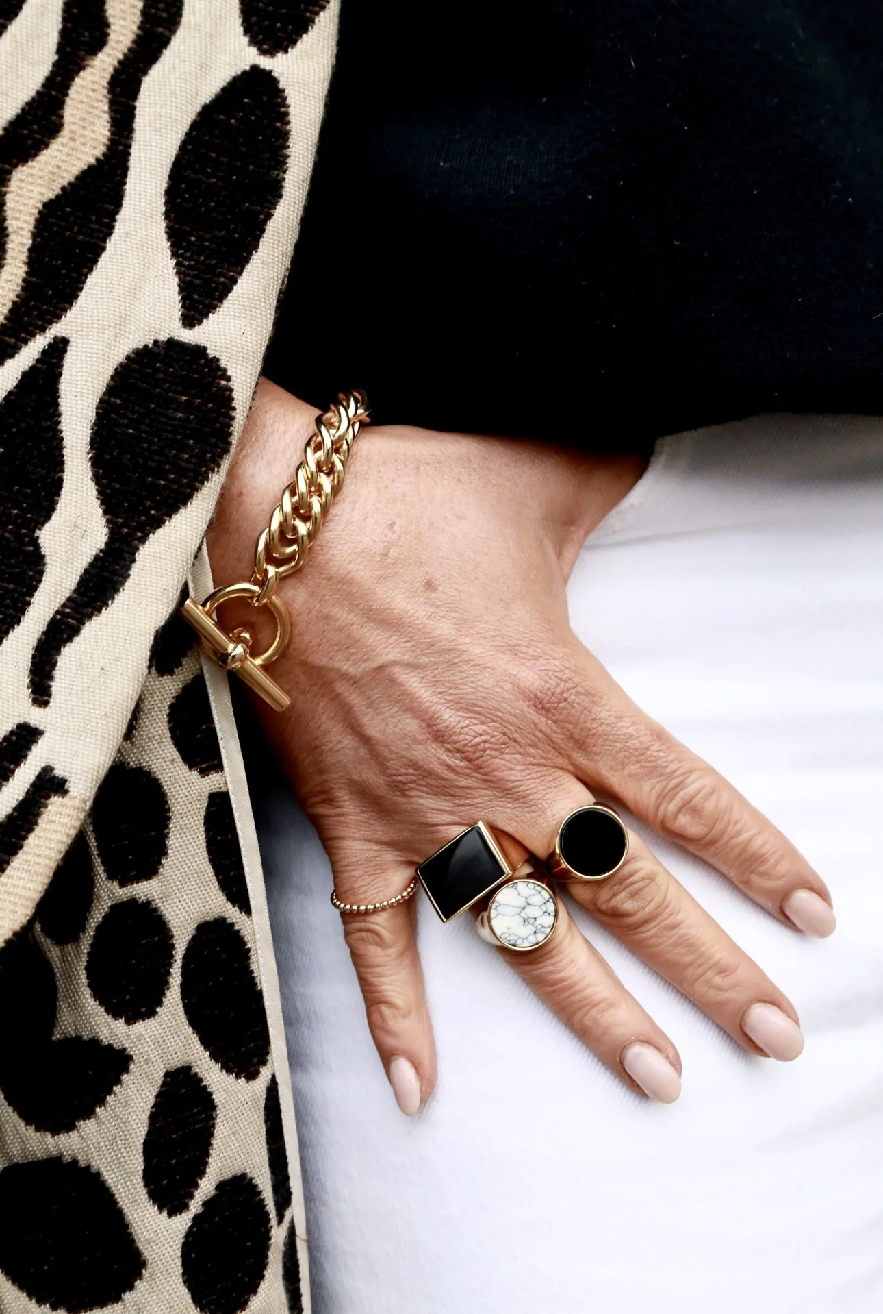 Close-up of a person's hand wearing rings and a bracelet, with a leopard-print fabric on the left and a black and white outfit and white pants.