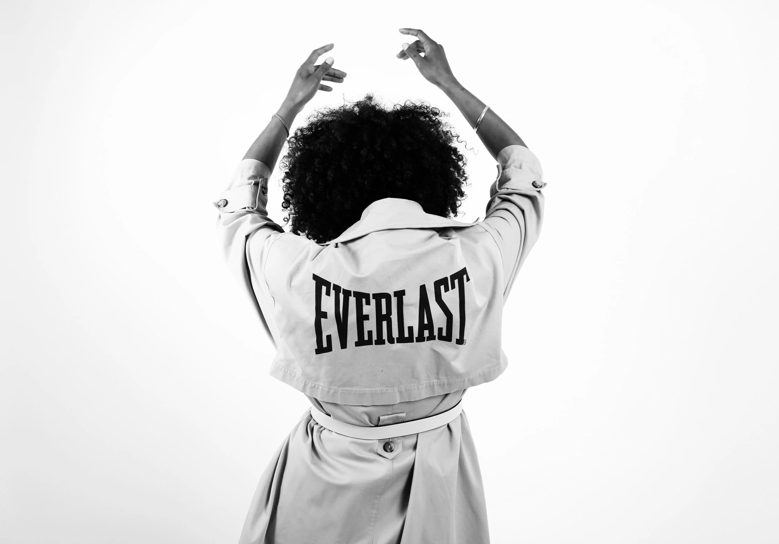 Back view of a woman with curly hair wearing a light-colored jacket with the word 'EVERLAST' on the back, raising her arms against a plain white background.
