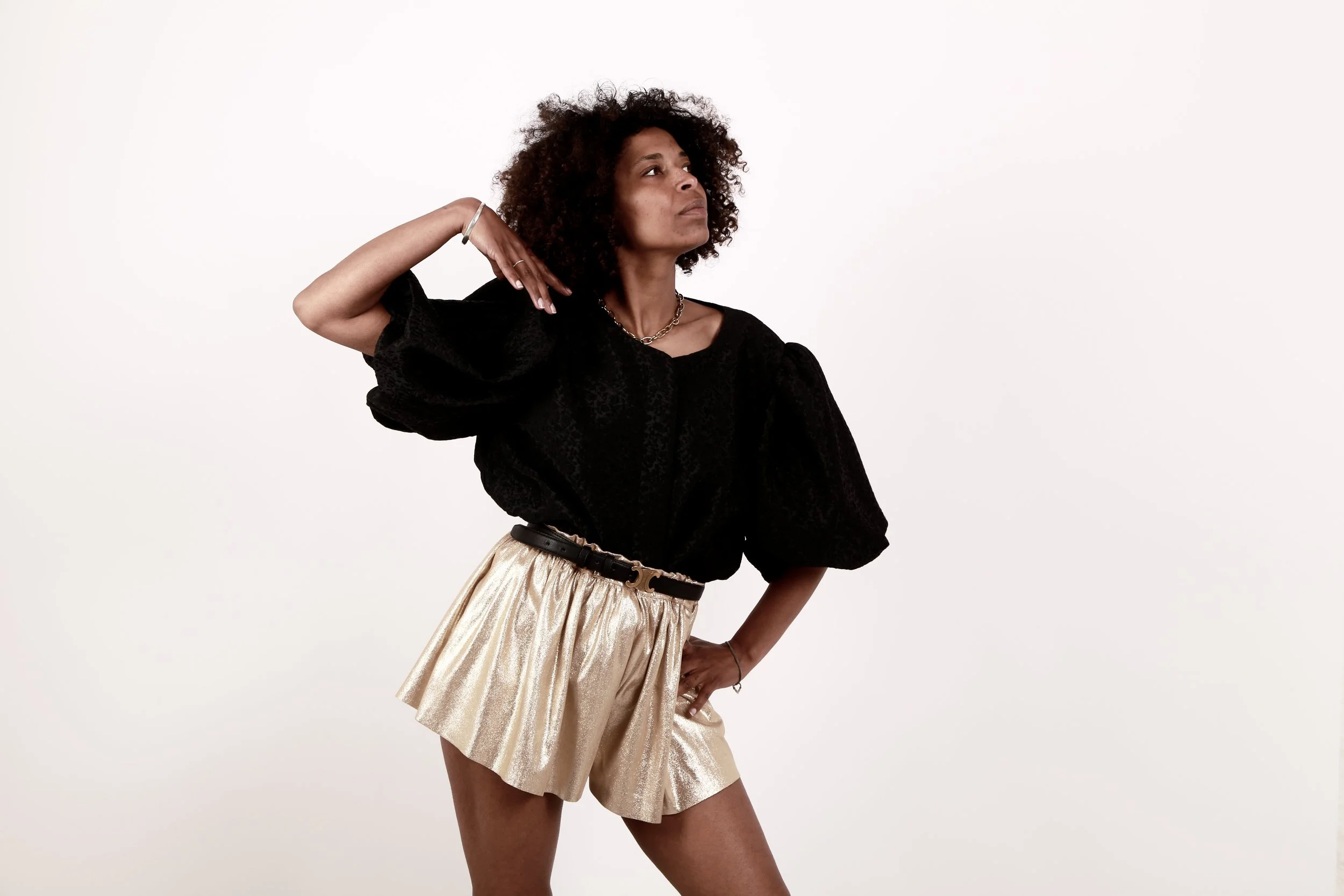 A woman with curly hair wearing a black puff-sleeve top, shiny gold shorts, and a black belt, posing with one hand on her hip and the other near her head against a plain white background.
