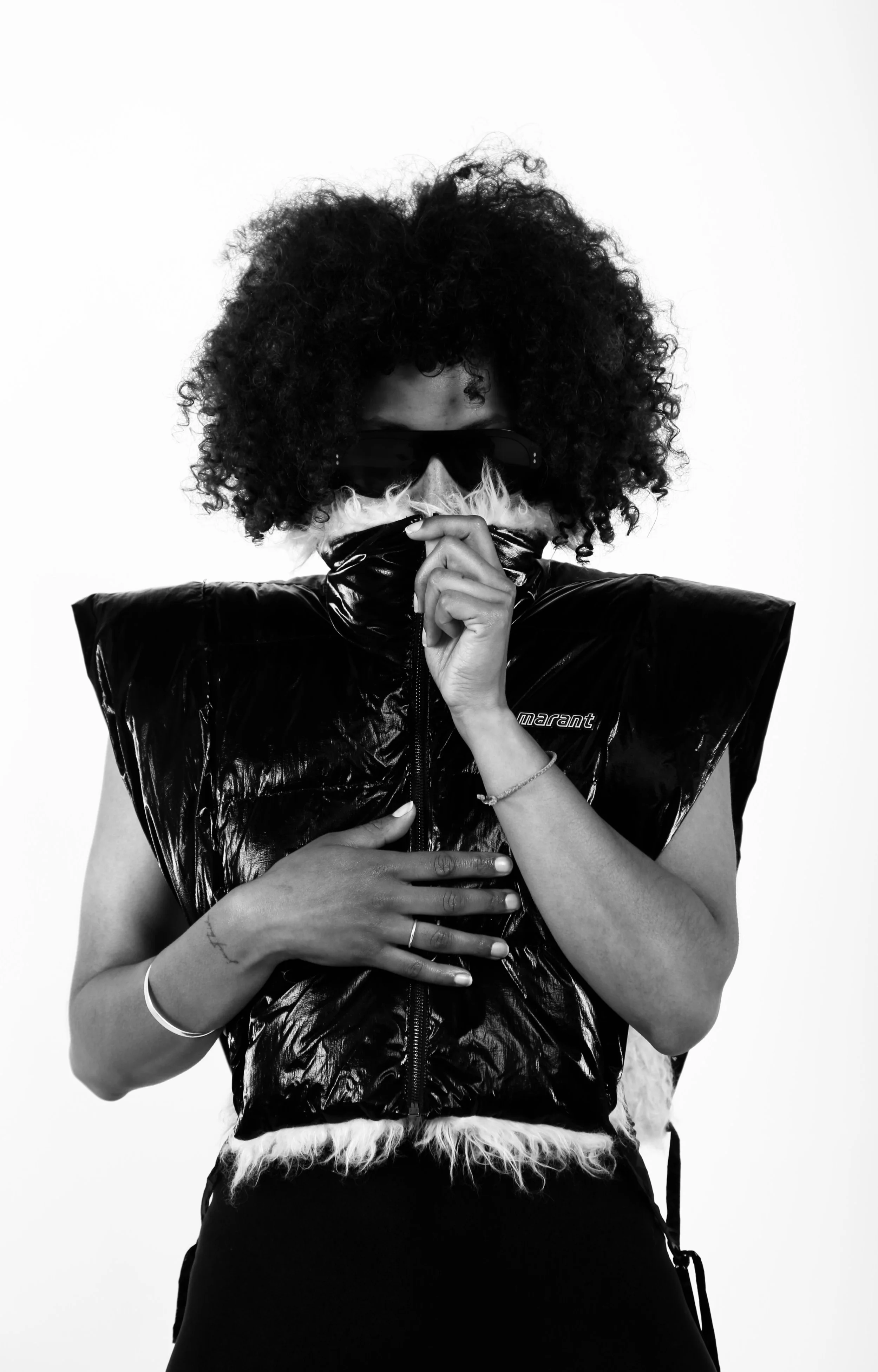 A person with curly hair wearing sunglasses, a shiny black sleeveless jacket, and a mask, holding their hand to their chest against a plain white background.