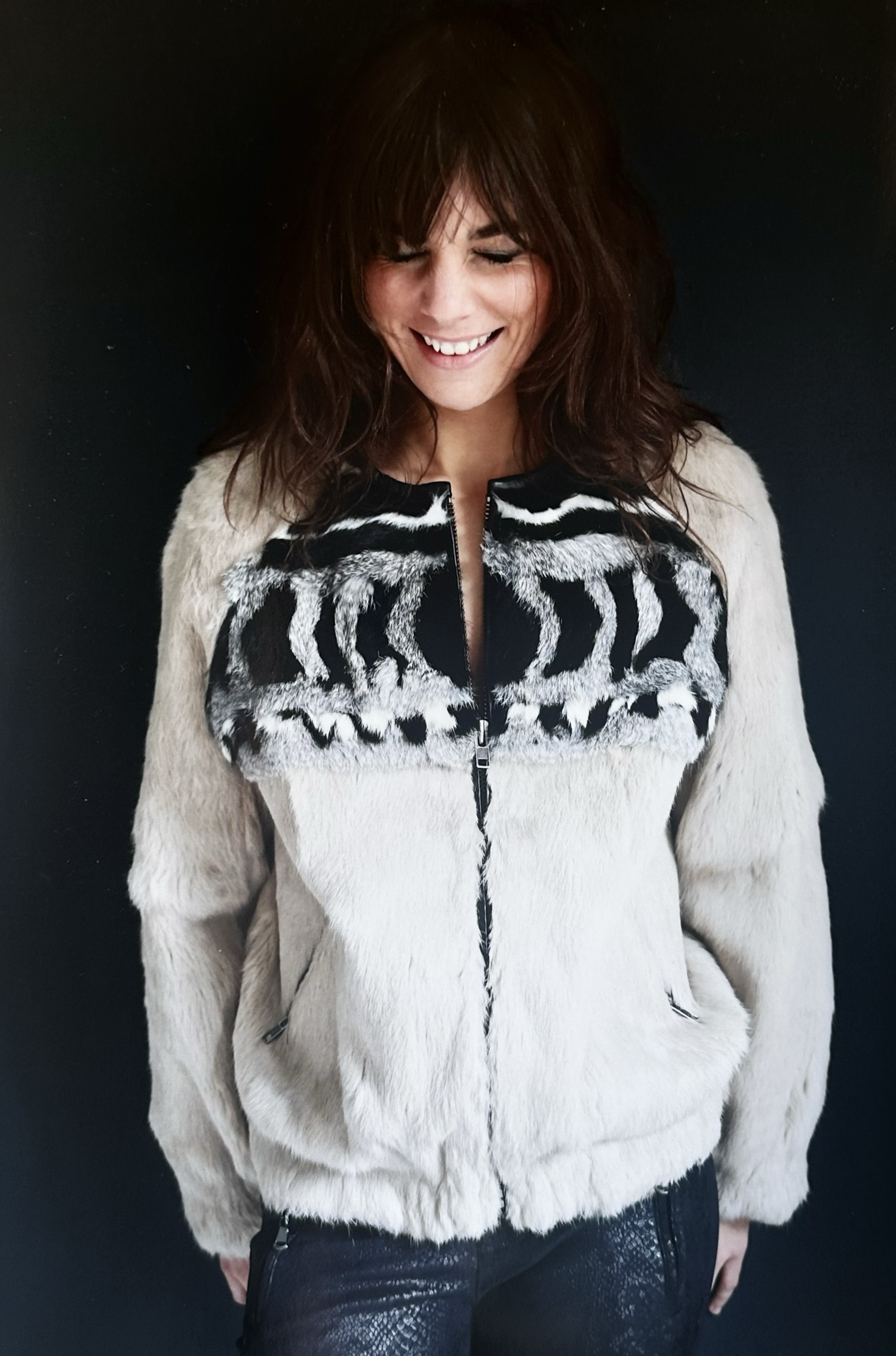 A woman with brown hair smiling, wearing a beige furry jacket with black and white patterned details on the chest and shoulders, standing against a dark background.