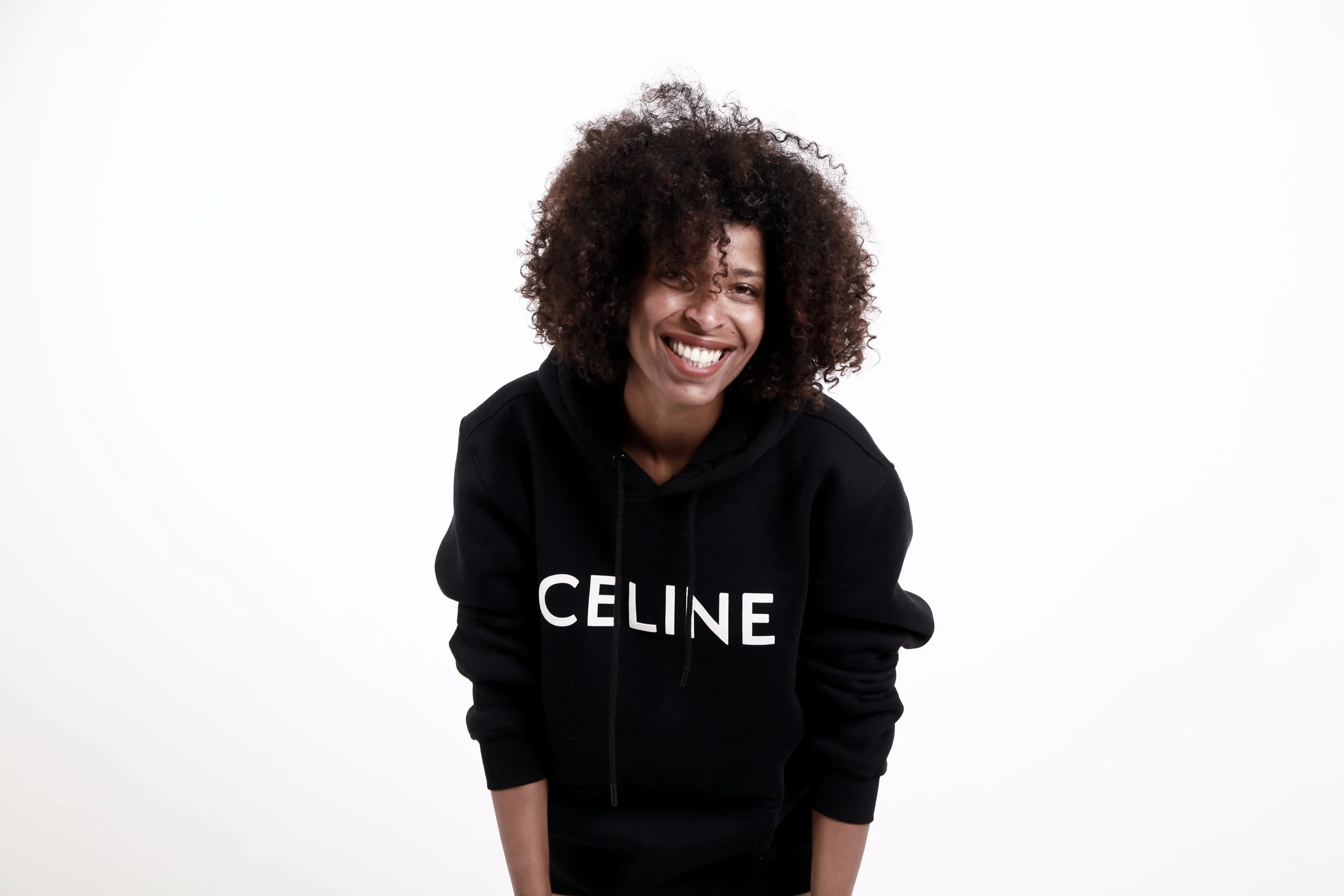 A woman with curly hair smiles and leans forward, wearing a black hoodie with the word 'CELINE' on it, against a plain white background.