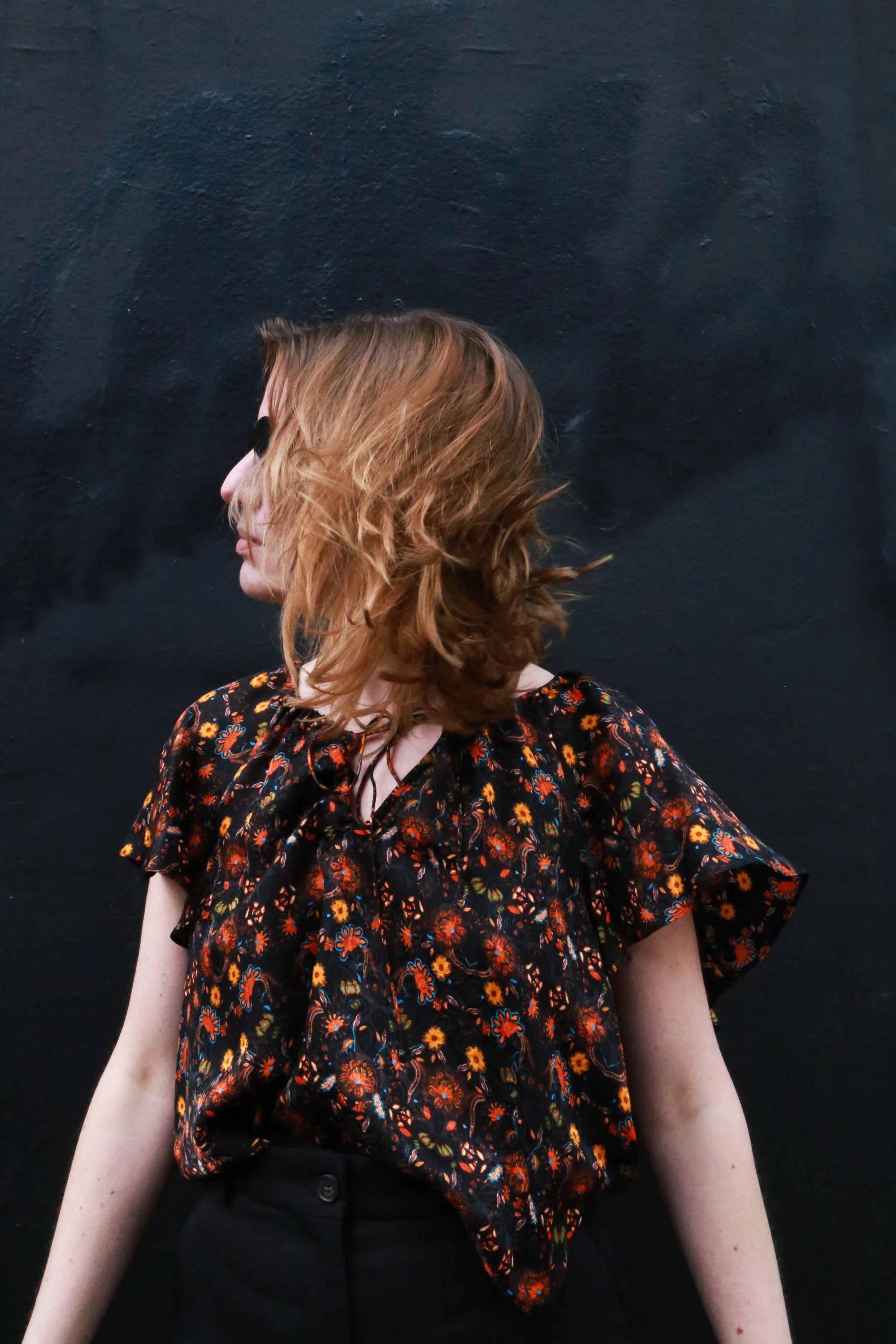 A woman with curly red hair wearing sunglasses, a black floral blouse, and black pants standing against a dark wall, looking to her left.