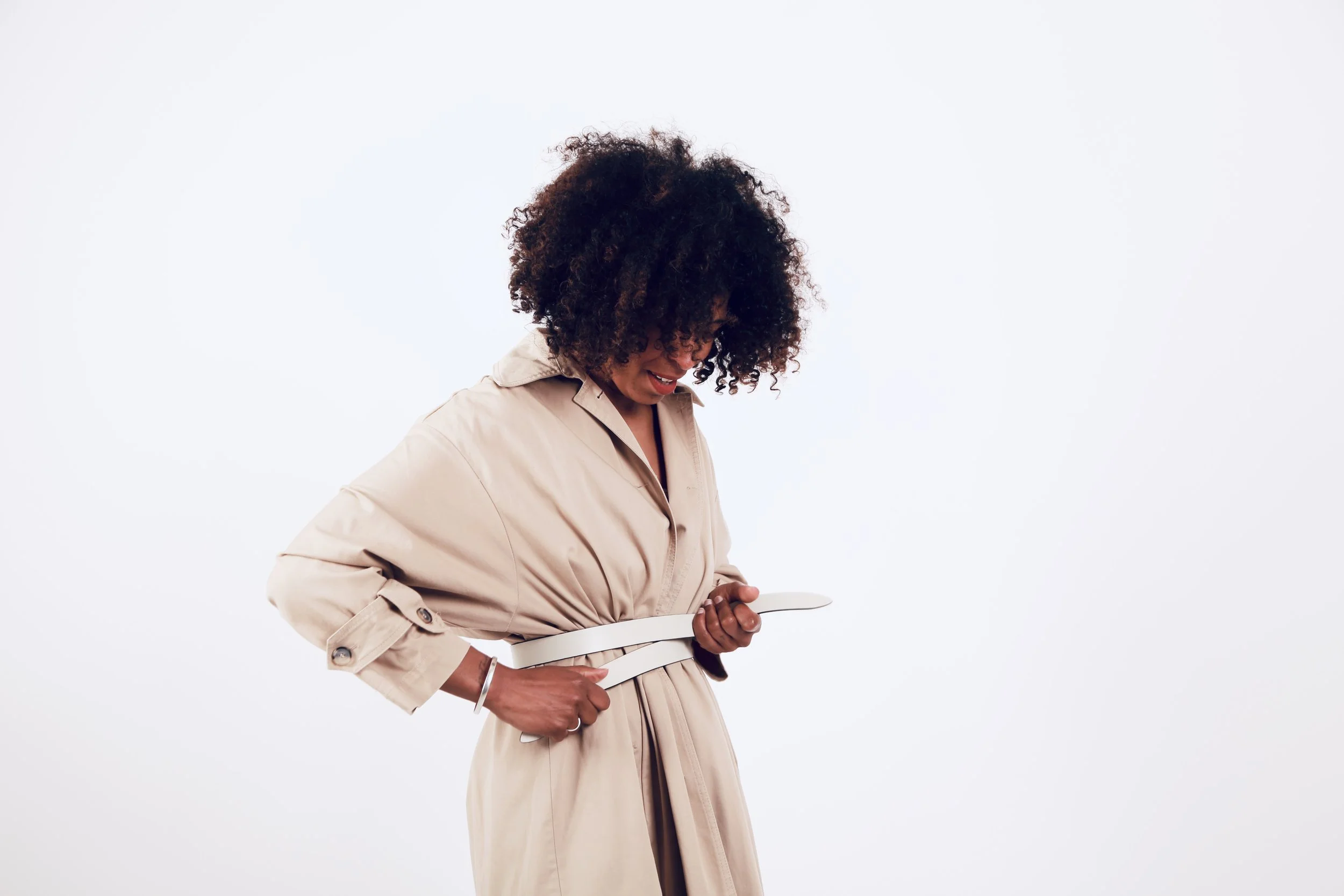 A woman in a beige trench coat looking down and smiling, adjusting a white belt at her waist against a plain white background.