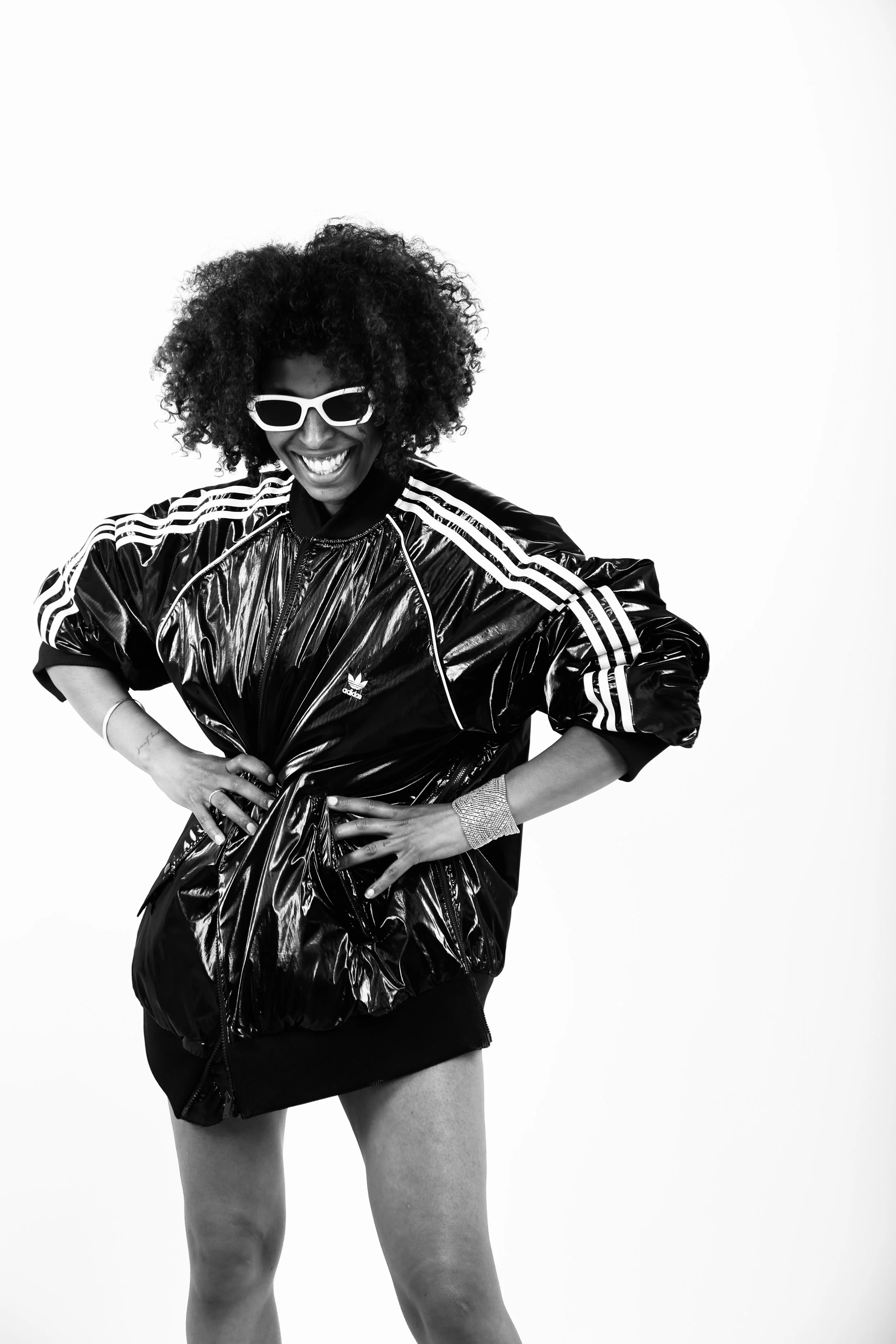 A woman with curly hair wearing sunglasses, a shiny black Adidas jacket, and a short skirt, posing with a smile against a plain background.