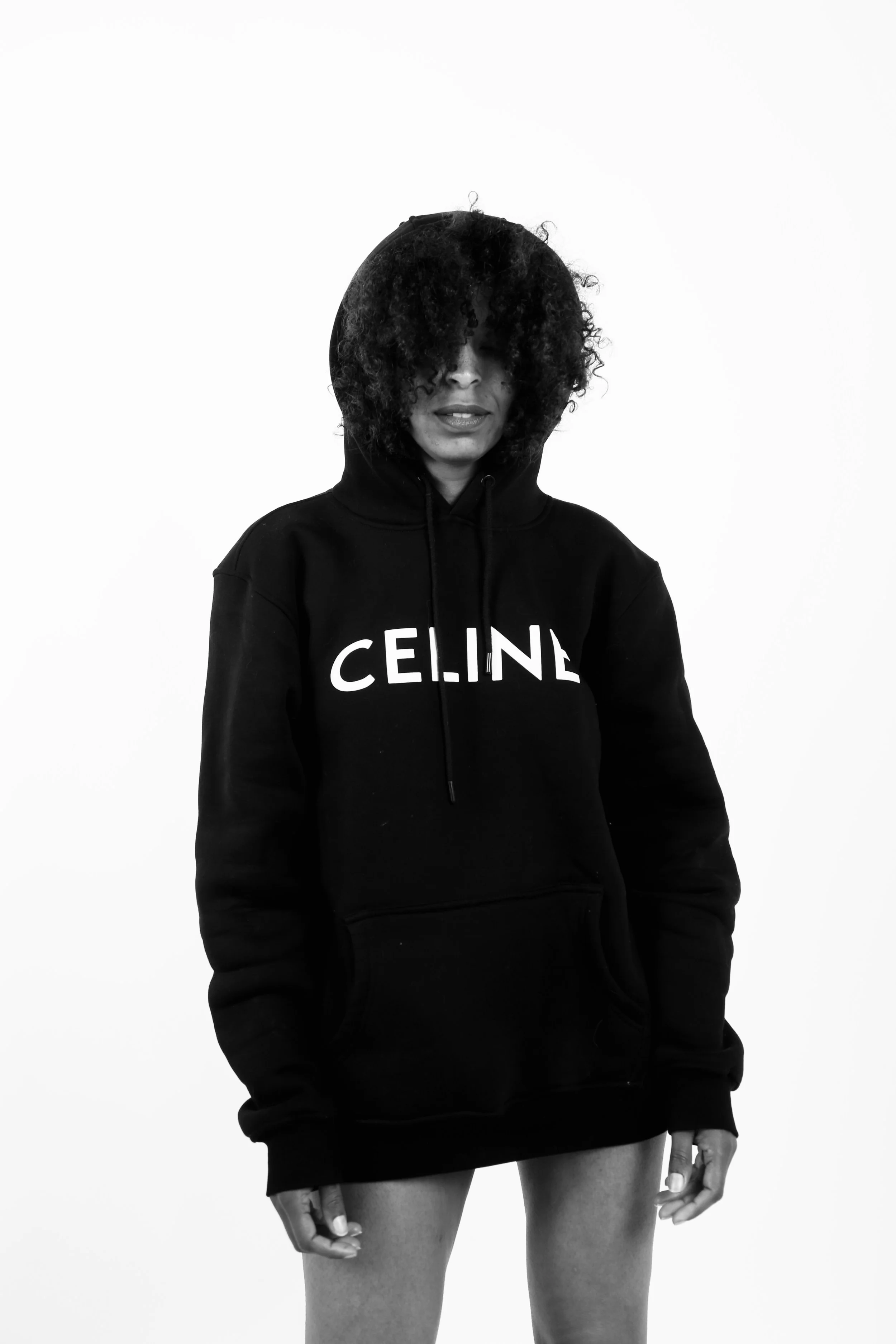Black and white photo of a woman wearing a black hoodie with the word 'CELINE' printed on it. She has curly hair partially covering her face, standing against a plain white background.