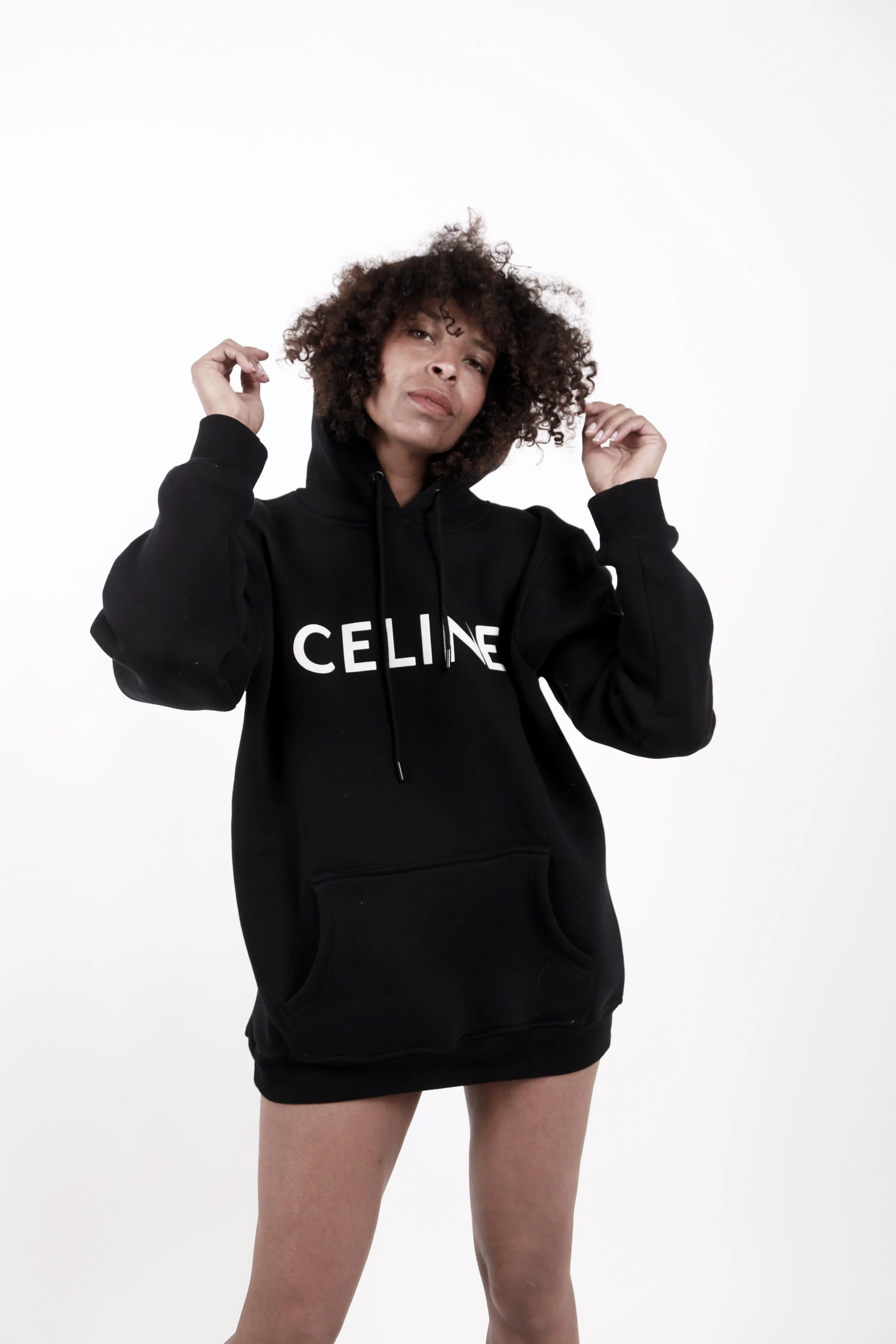 Woman with curly hair wearing a black hoodie with 'CELINE' written on it, standing against a white background.