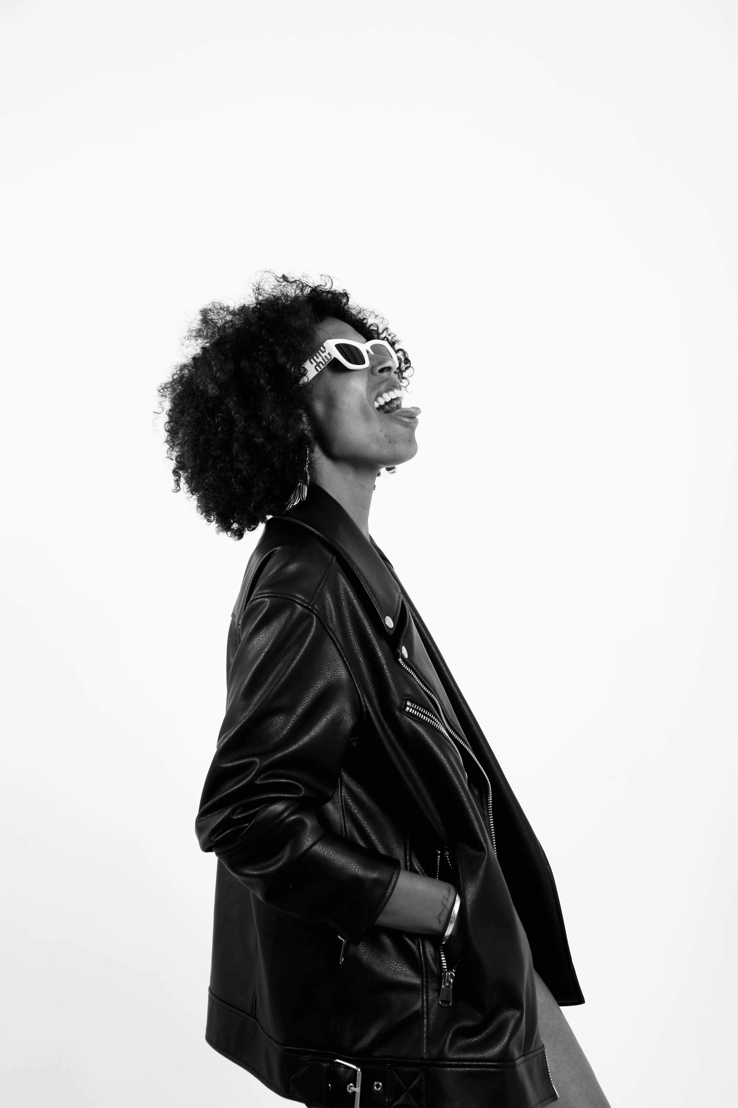 A woman with curly hair wearing sunglasses and a black leather jacket, laughing and sticking out her tongue, against a plain white background.