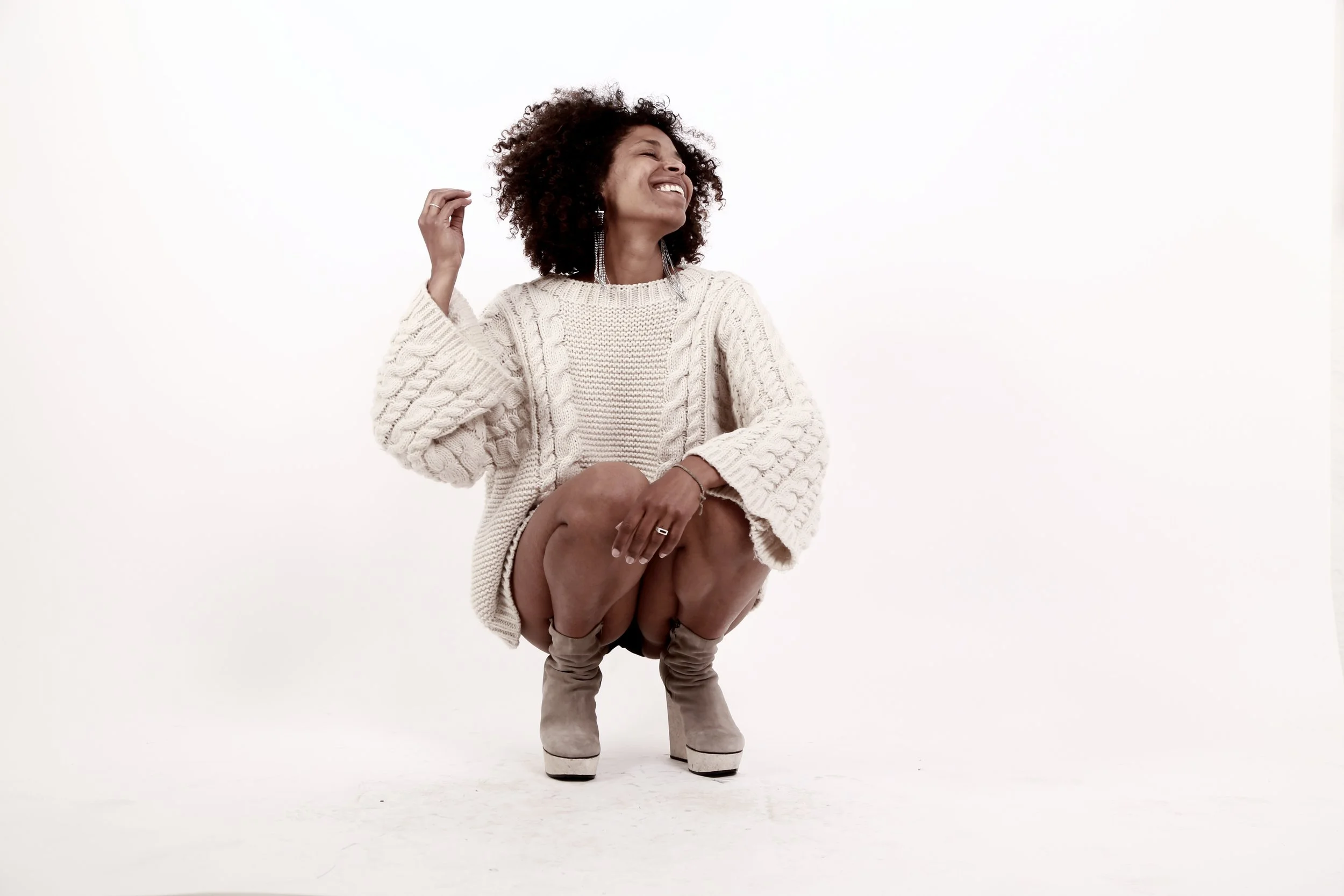 A woman in a cream cable-knit sweater and beige high-heeled boots squats on the floor, smiling and looking upward against a plain white background.