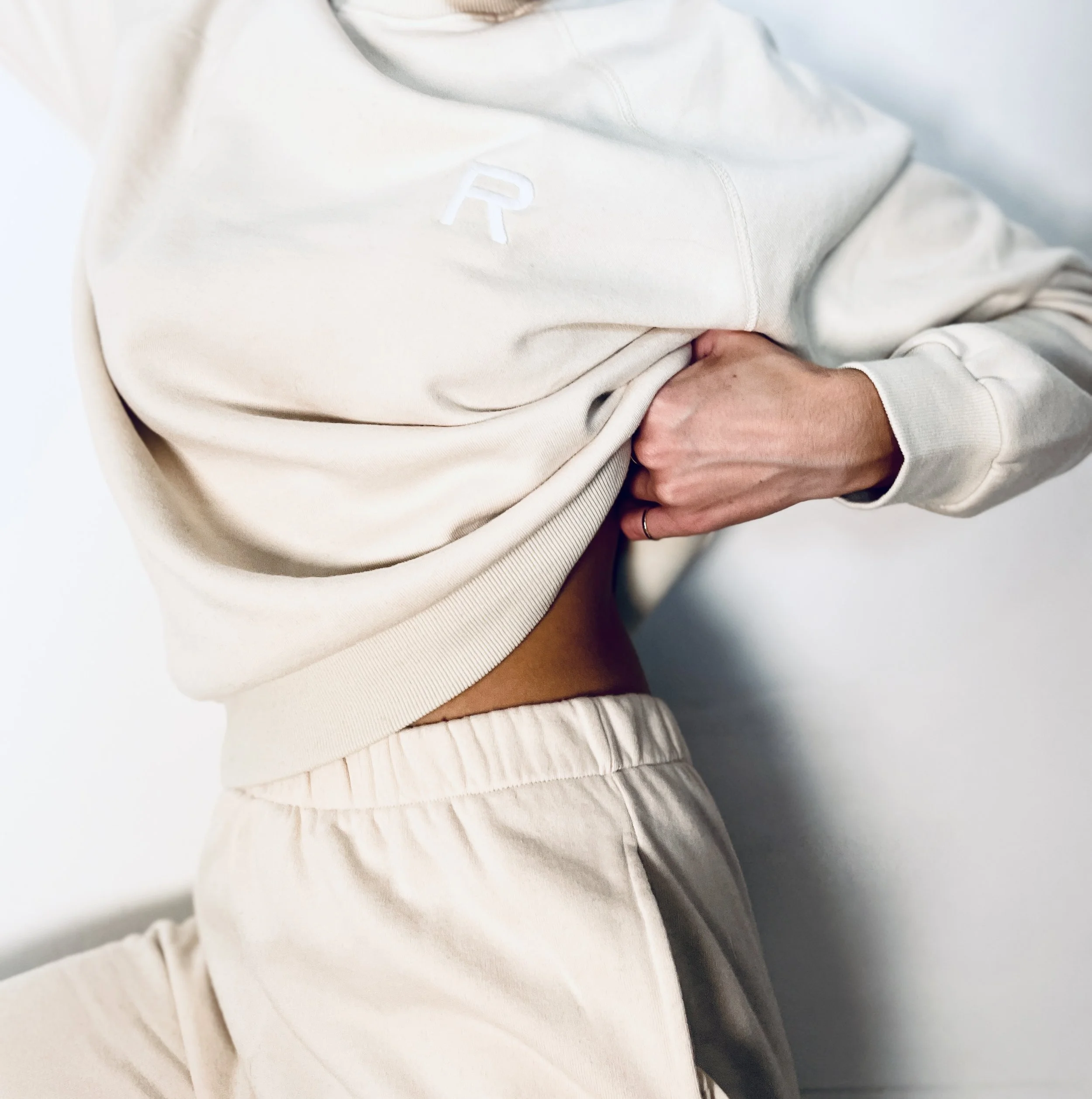 Person lifting their sweatshirt to reveal their abdomen, wearing beige sweatpants and a cream-colored sweatshirt with a small logo.